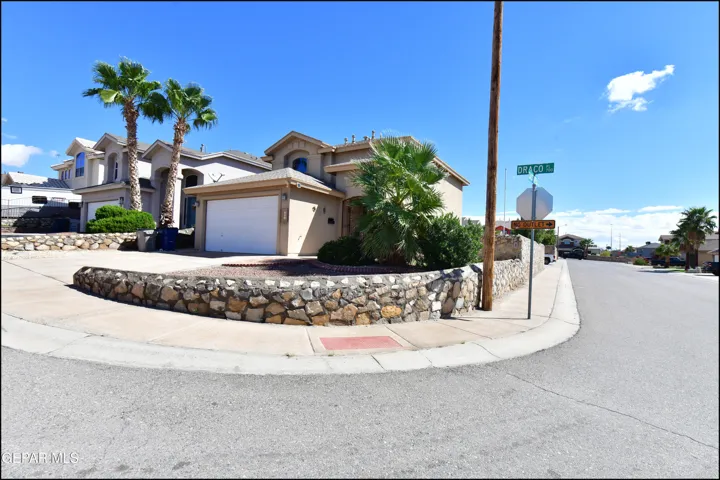 Stucco exterior with an attached garage, featuring a stone retaining wall and mature palm trees