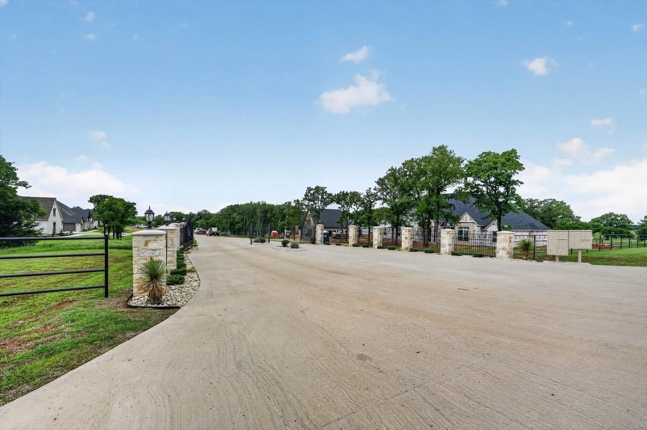 Concrete driveway leading to an entry featuring stone pillars and black metal fencing