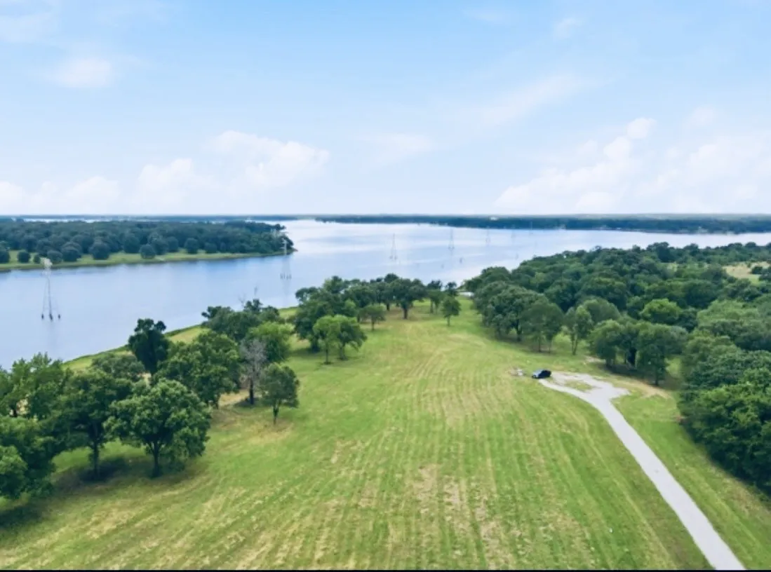 Expansive waterfront property featuring a wide body of water, lush green fields, mature trees, and a paved private drive