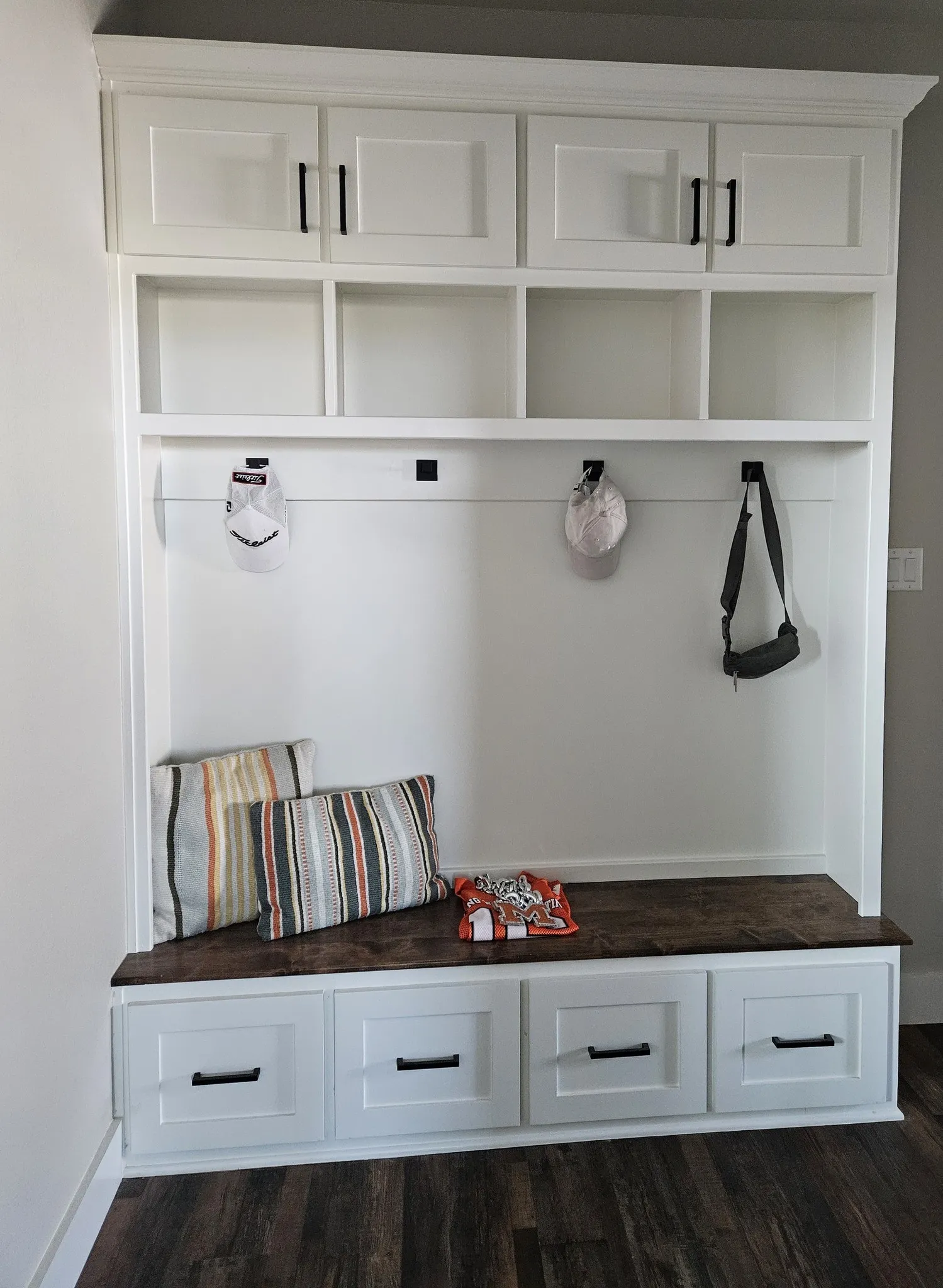 Example Photos: Built-in mudroom storage featuring white shaker cabinetry, a wood-finish bench, integrated cubbies, and black hardware