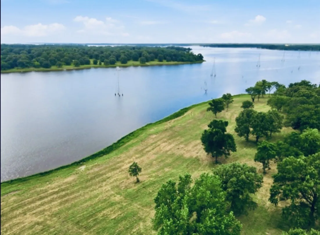 Expansive lakefront property featuring a gently sloping grassy terrain, numerous mature trees, and direct water access