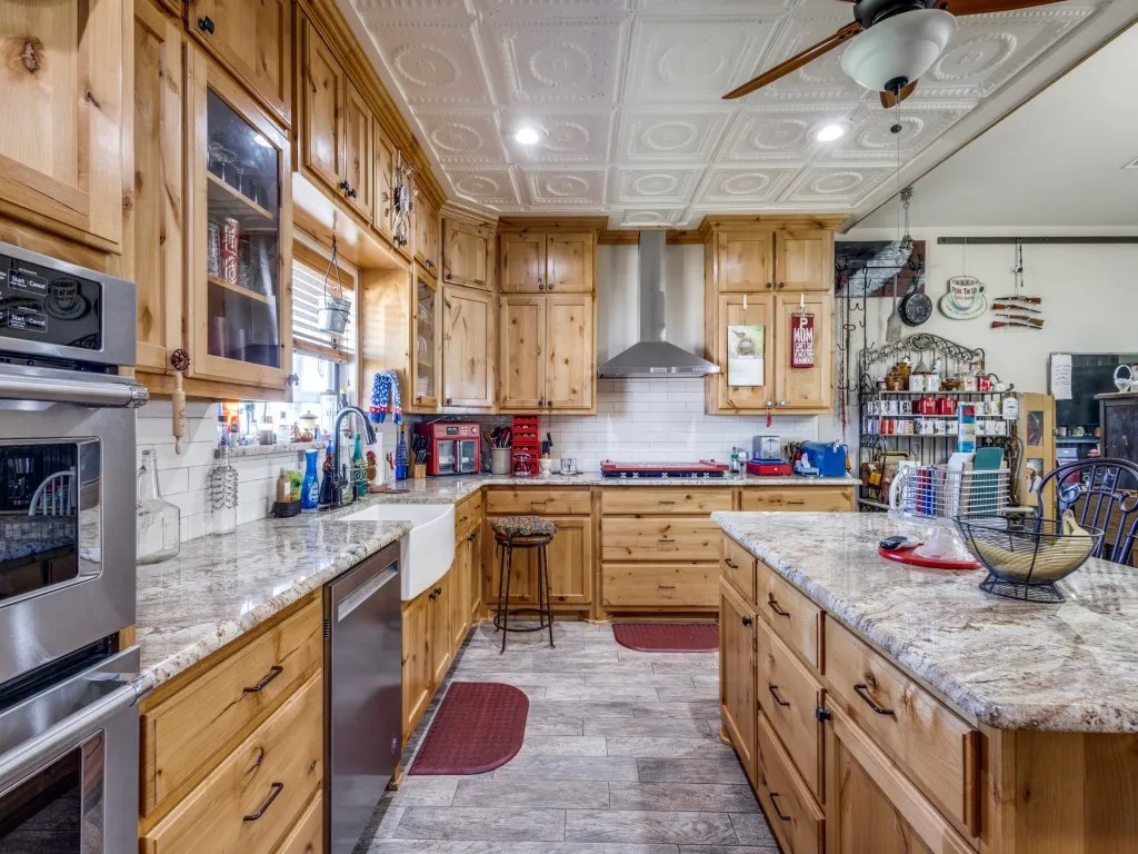 Kitchen featuring ceiling fan, wall chimney range hood, and light stone counters