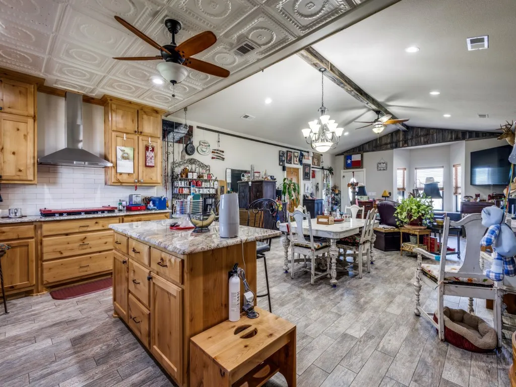 Kitchen featuring wall chimney exhaust hood, ceiling fan with notable chandelier, light stone counters, a kitchen island, and tasteful backsplash