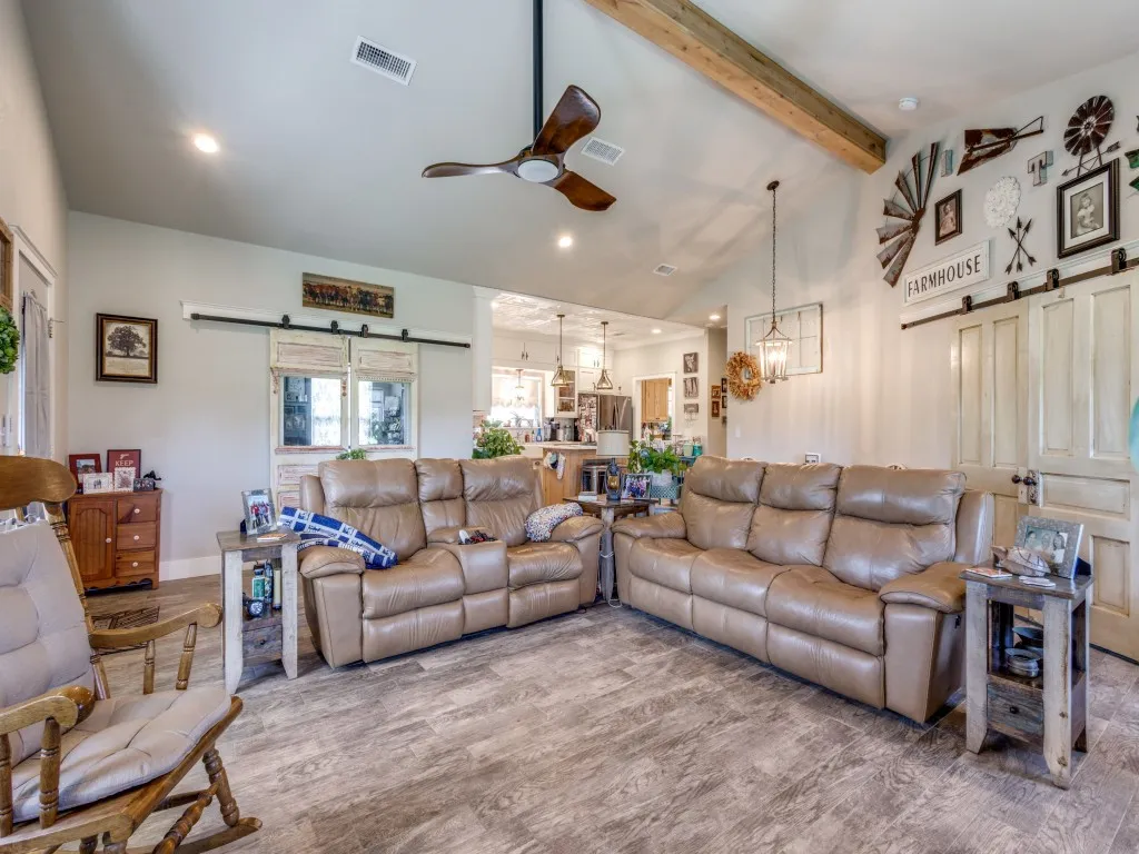 Living room with high vaulted ceiling, a barn door, ceiling fan with notable chandelier, beam ceiling, and hardwood floors