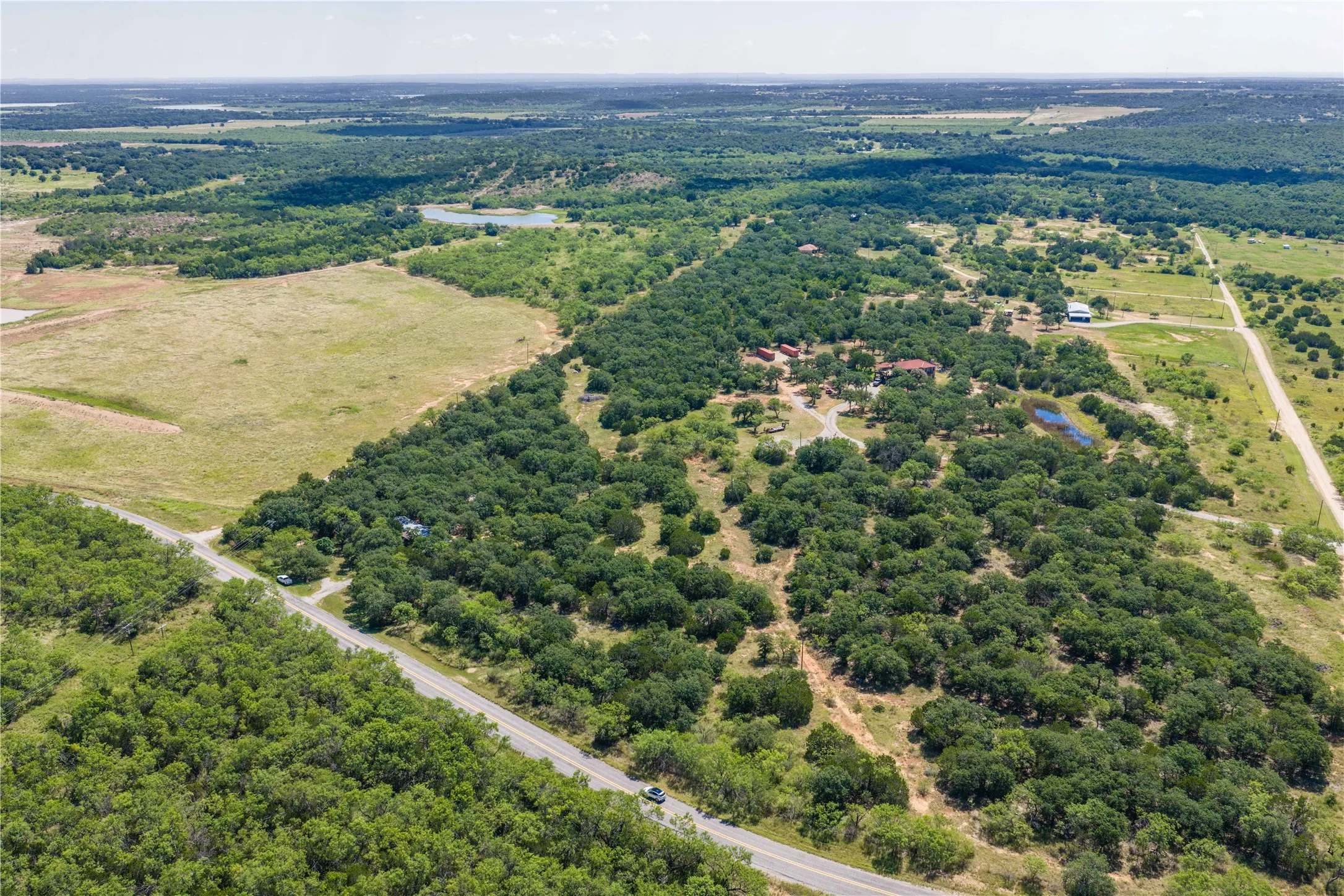 Expansive rural property featuring a paved road frontage, extensive tree coverage, multiple structures, open fields, and a visible pond
