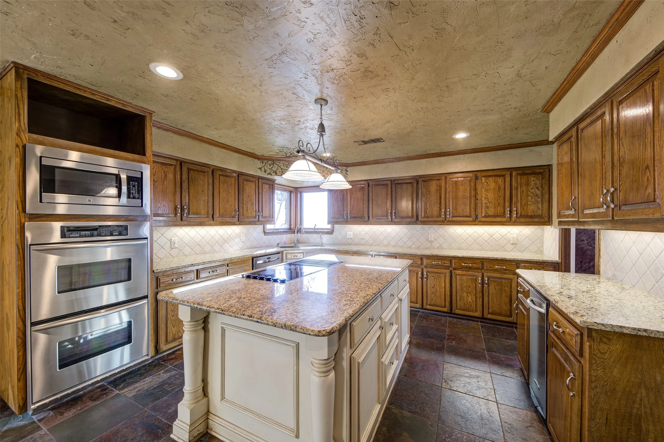 Kitchen with stainless steel appliances, stone tile floors, ornamental molding, brown cabinetry, and light stone counters