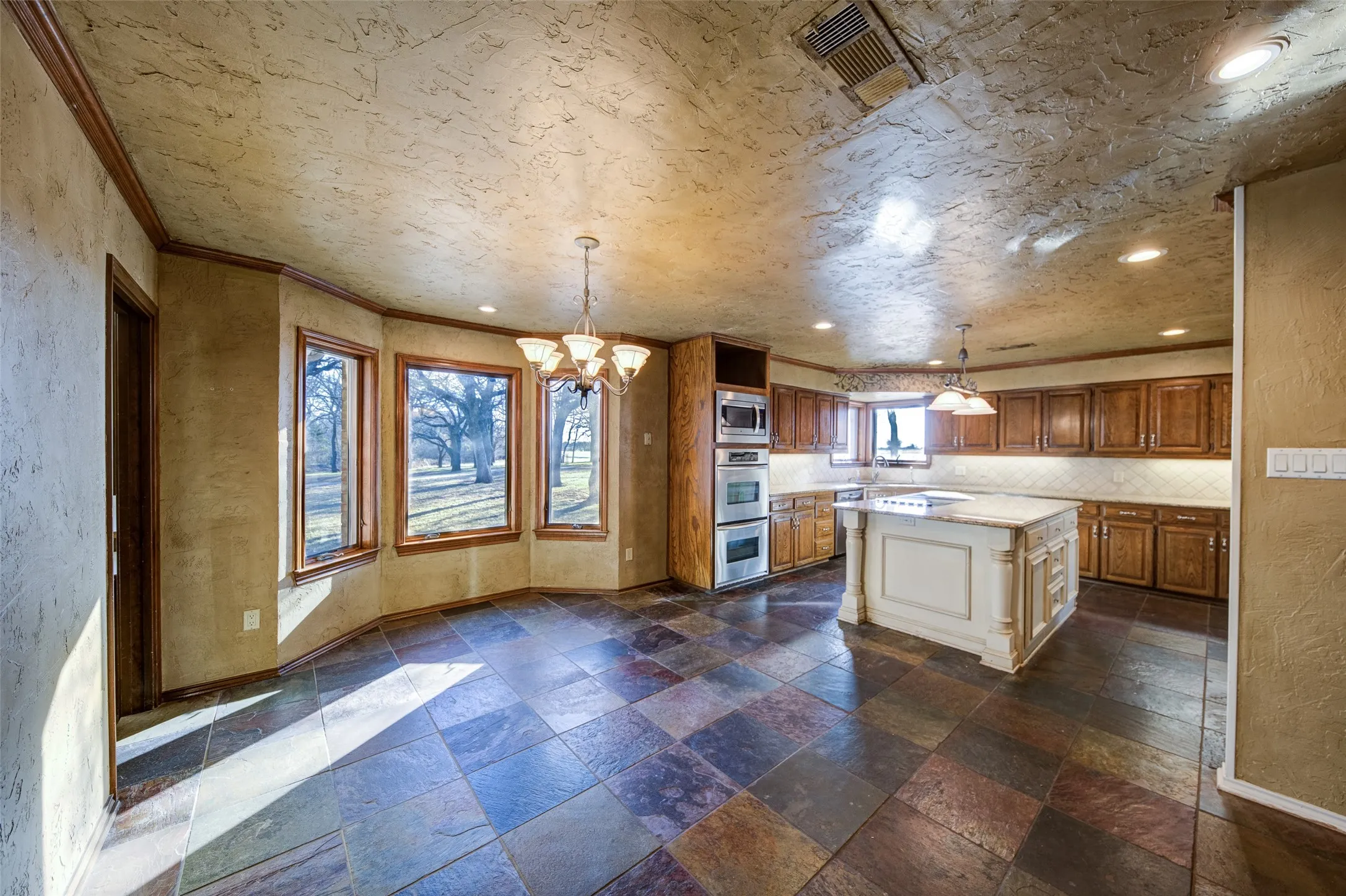 Kitchen featuring appliances with stainless steel finishes, a chandelier, brown cabinetry, stone tile flooring, and decorative backsplash