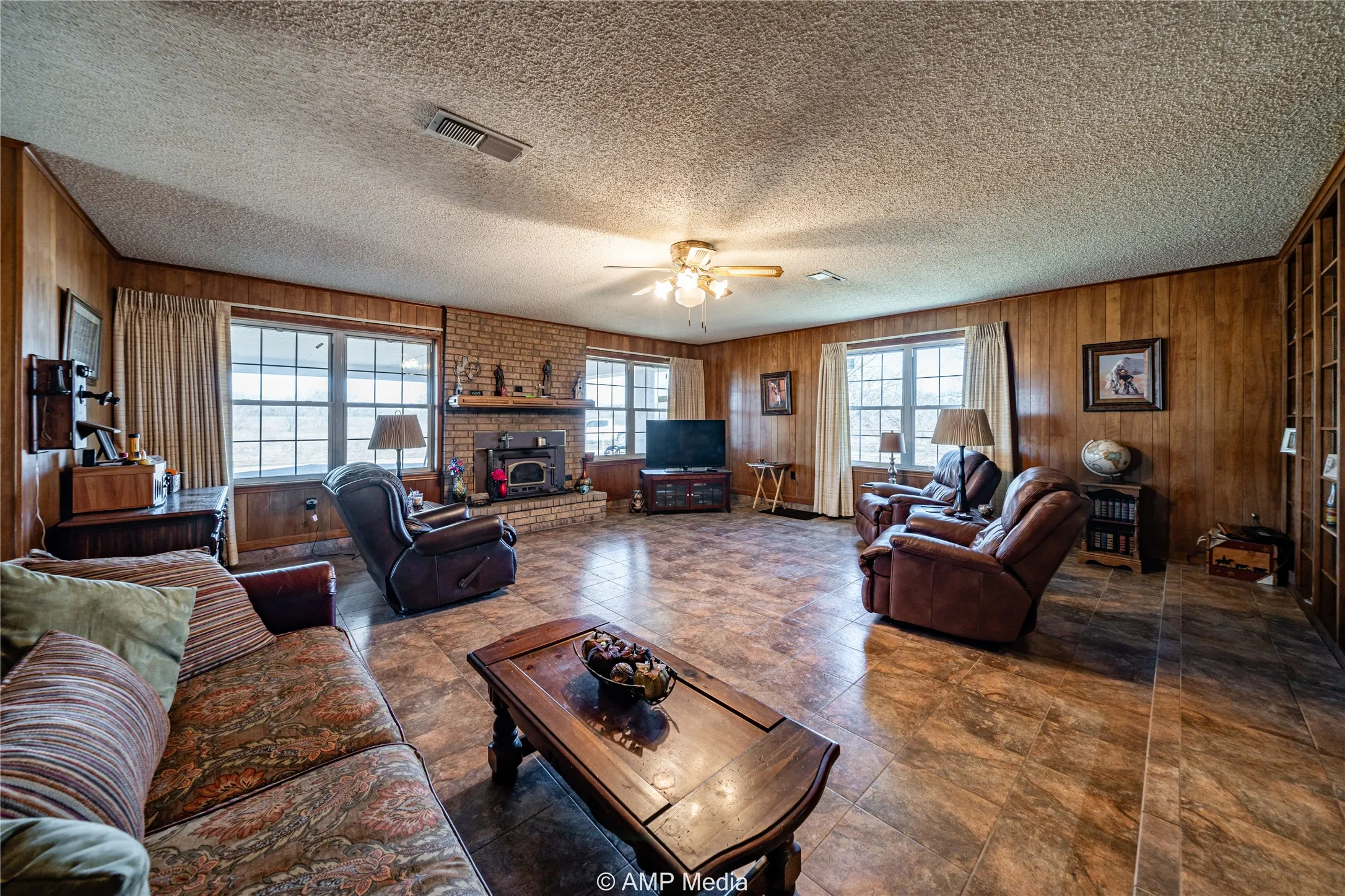 Living room featuring a brick fireplace, a textured ceiling, ceiling fan, and wood walls