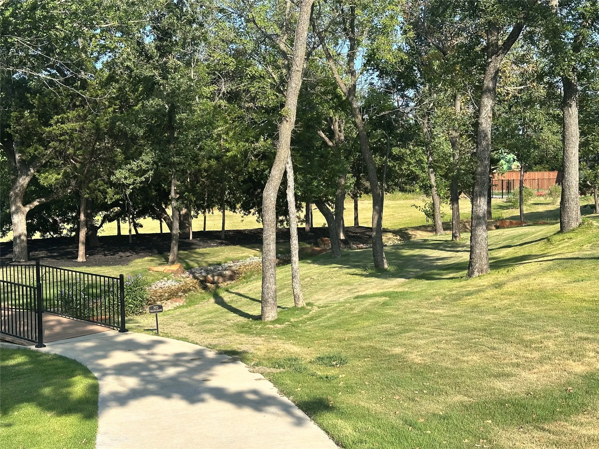 Expansive green space featuring mature trees, a paved walkway, a black metal bridge, and a natural creek bed
