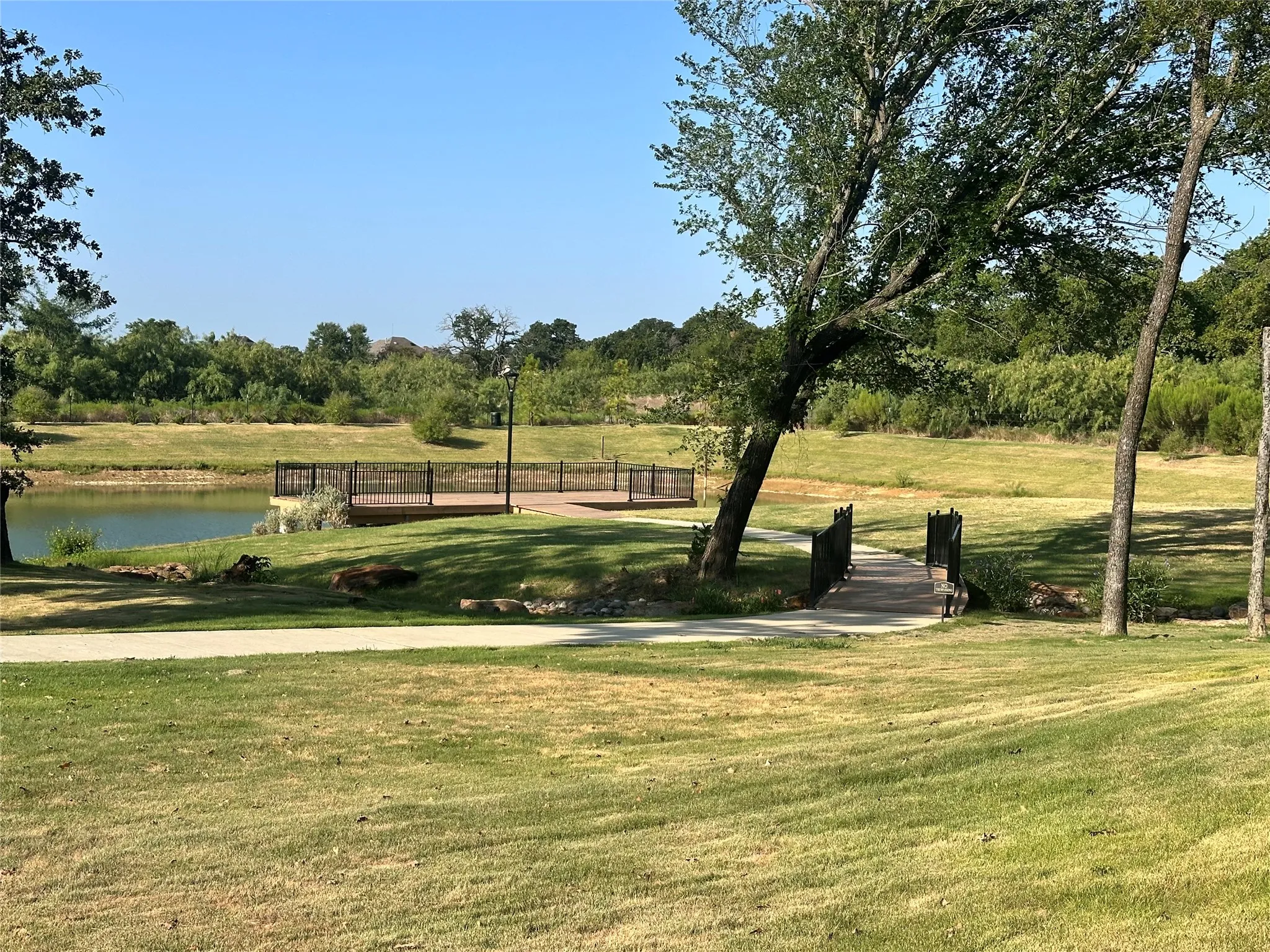 Waterfront decking with black metal railing, concrete pathway, and wooden bridge, all surrounded by green landscaping
