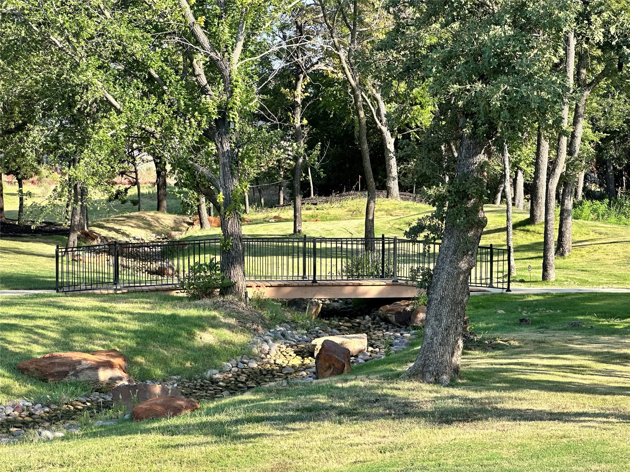 Landscaped green space featuring a pedestrian bridge with black railings, a stone-lined creek bed, and mature trees