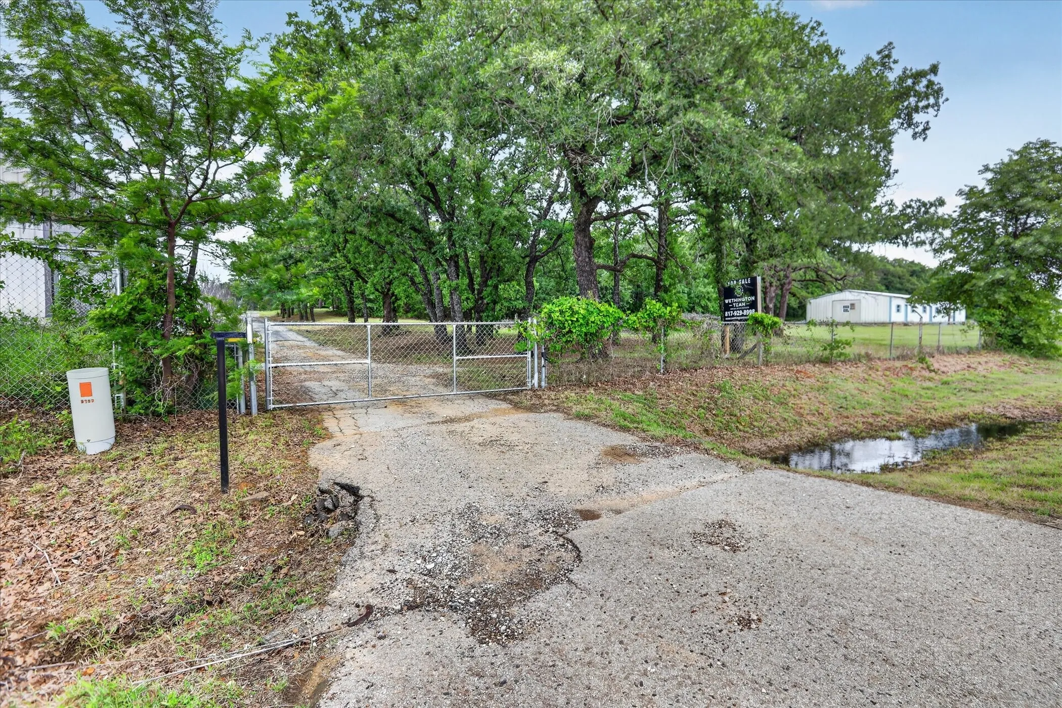 Gated entry with a gravel driveway, chain-link fencing, and mature trees