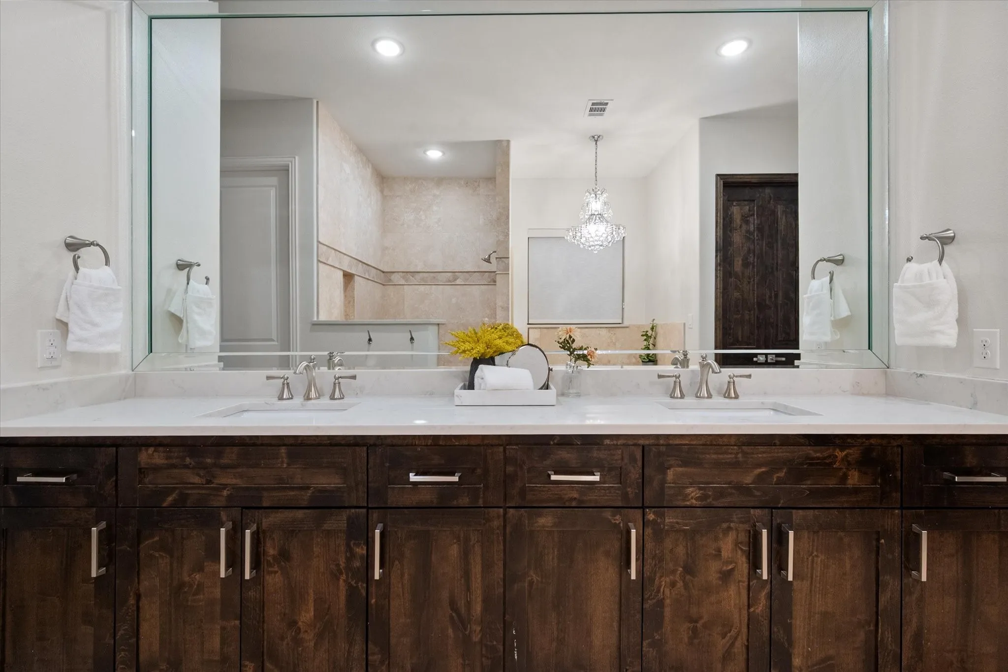 The primary bathroom features dual vanities with quartz counters, rich cabinetry, a large framed mirror and a sparkling chandelier over the soaking tub.