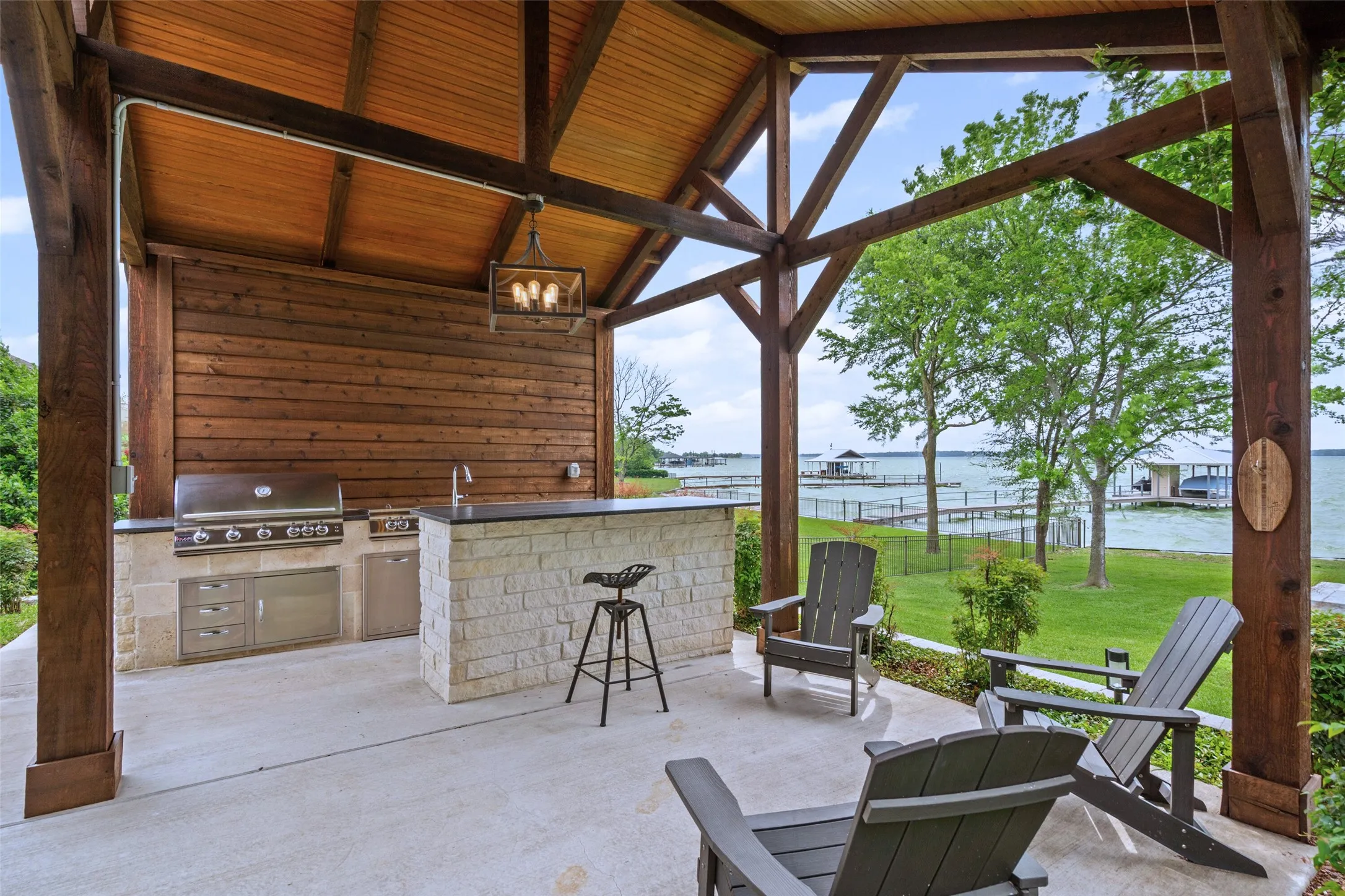 The fully equipped outdoor kitchen, complete with built-in grill and bar seating, sits beneath a vaulted pergola overlooking the lake.