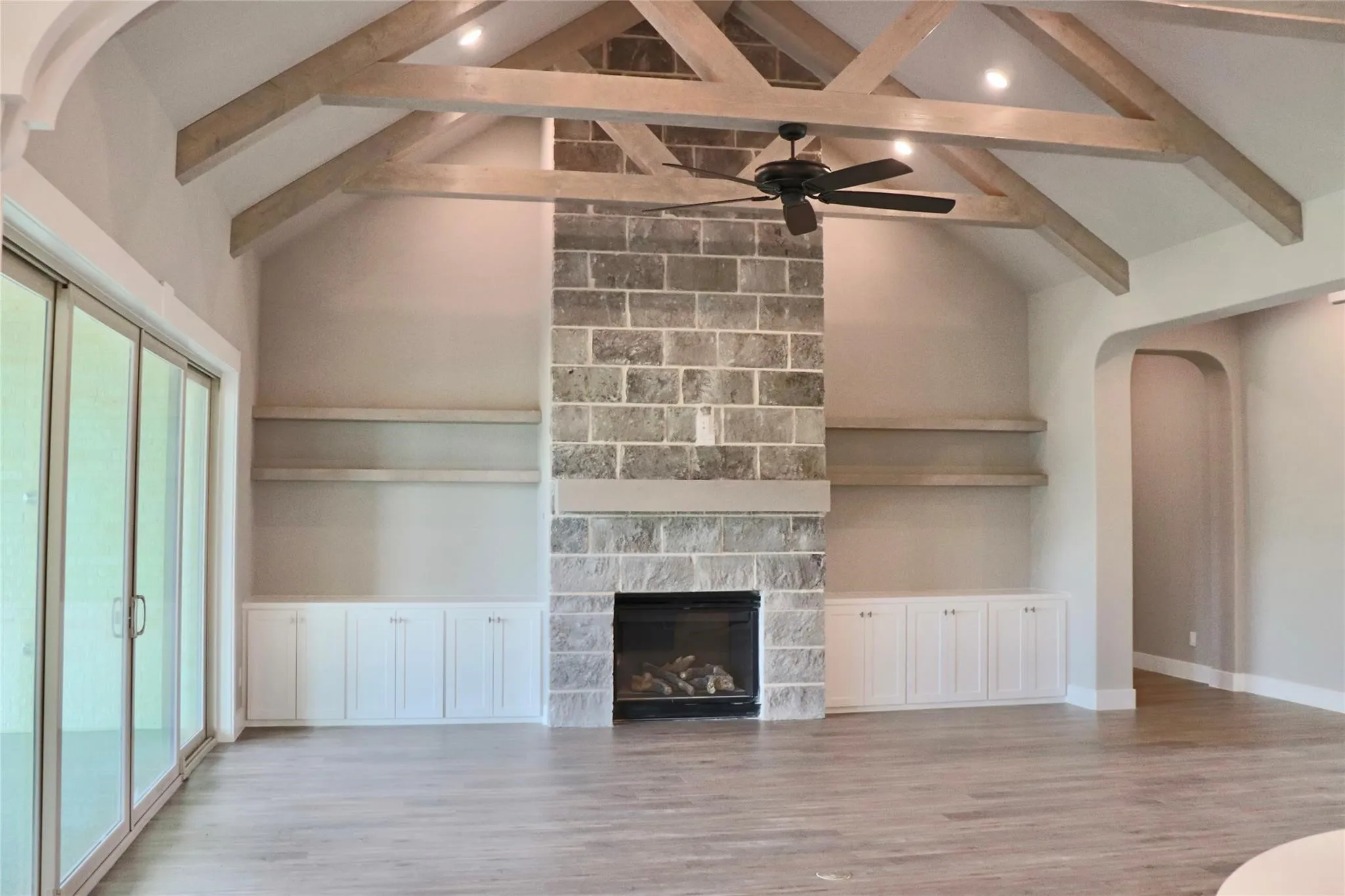 Unfurnished living room with beam ceiling, plenty of natural light, ceiling fan, wood finished floors, and a stone fireplace