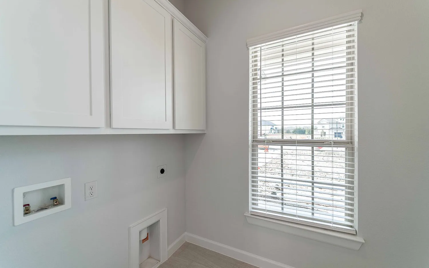 White shaker-style cabinetry, a single window with horizontal blinds, wood-finish flooring, and utility hookups