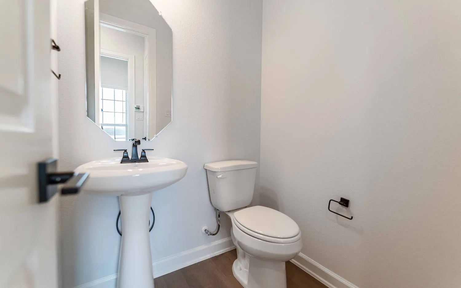 Pedestal sink with a matte black faucet, an octagonal wall-mounted mirror, and wood-finish flooring