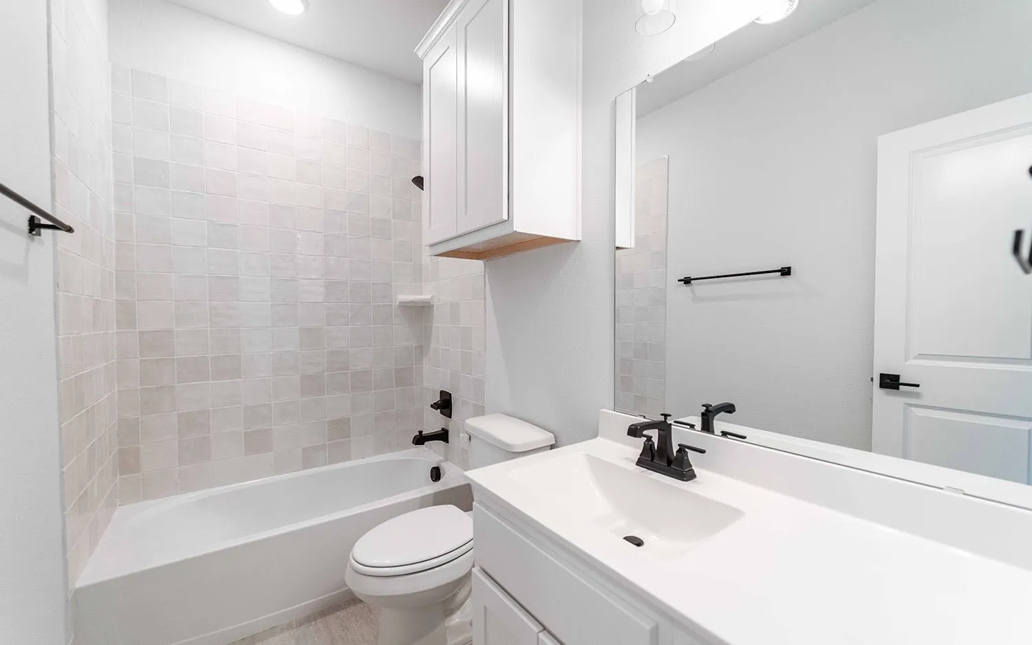 White subway tile shower surround, built-in bathtub, white vanity with integrated sink, matte black faucet, and overhead cabinetry