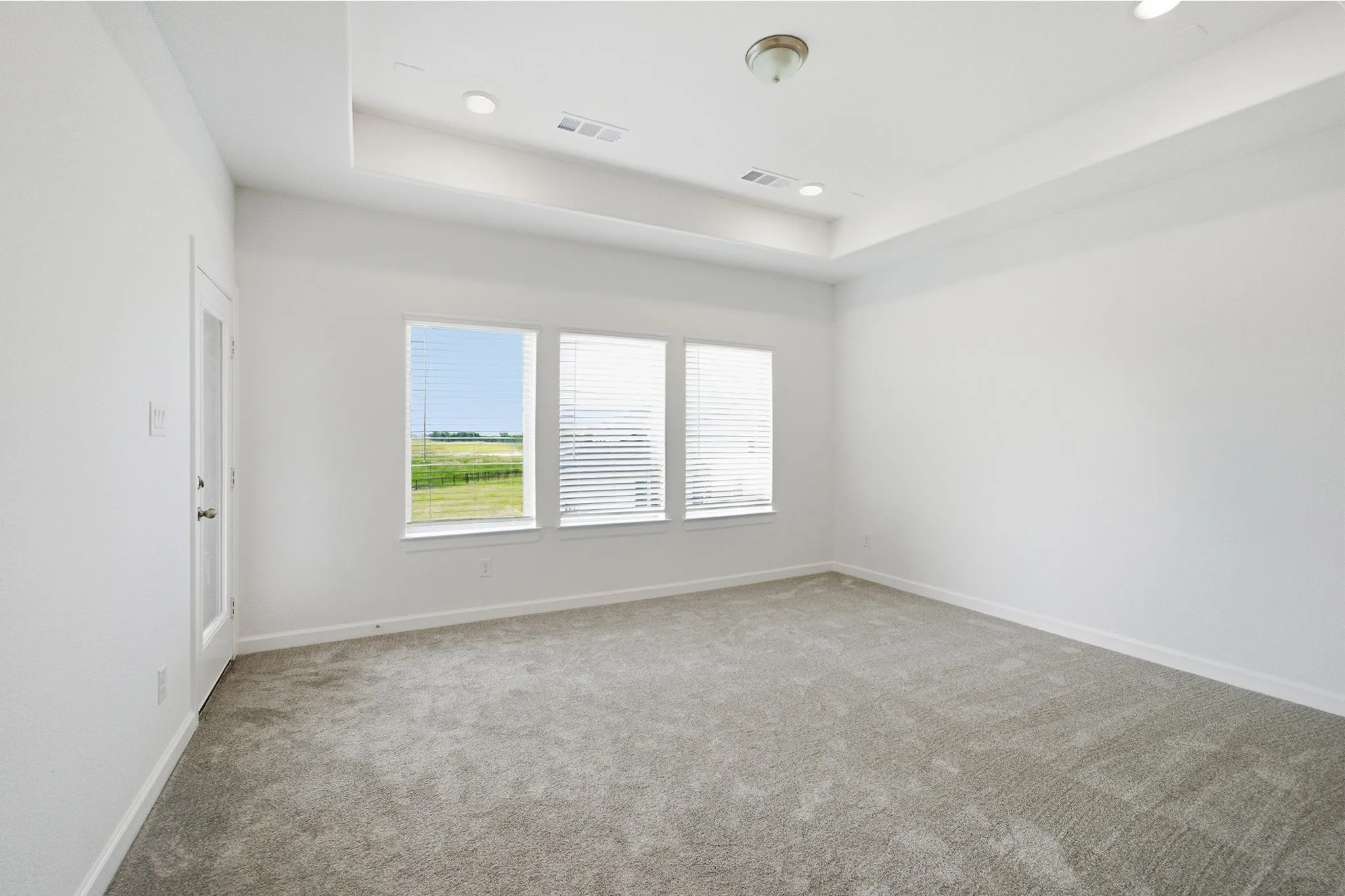 Spacious room featuring light gray carpet, white walls, a tray ceiling with recessed lighting, and three windows overlooking a green landscape