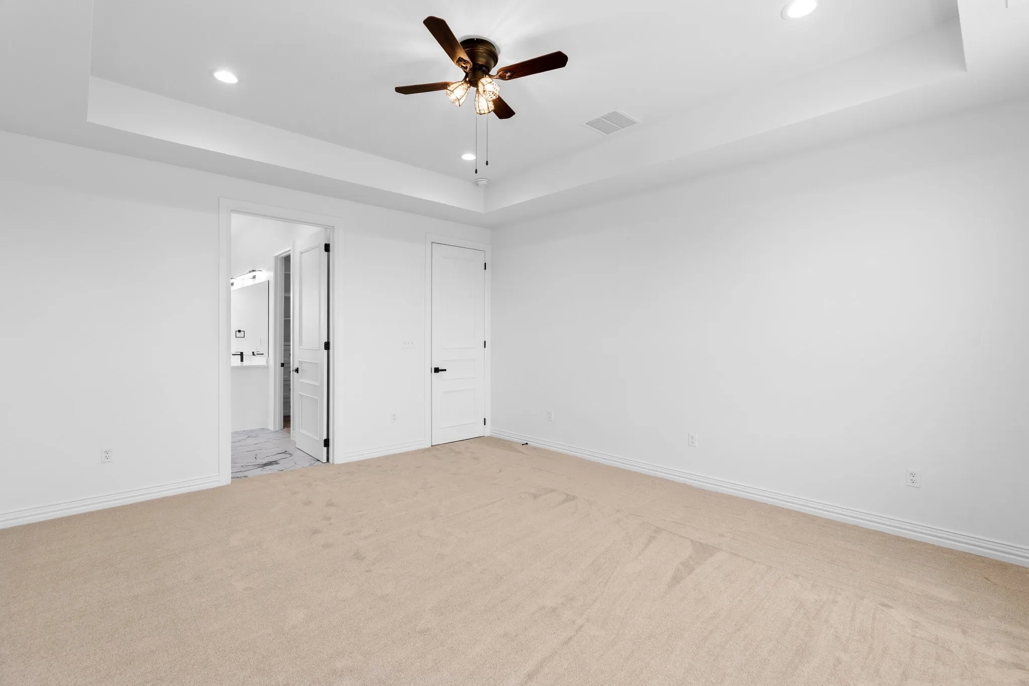 Unfurnished bedroom featuring a raised ceiling, light colored carpet, recessed lighting, ensuite bath, and ceiling fan