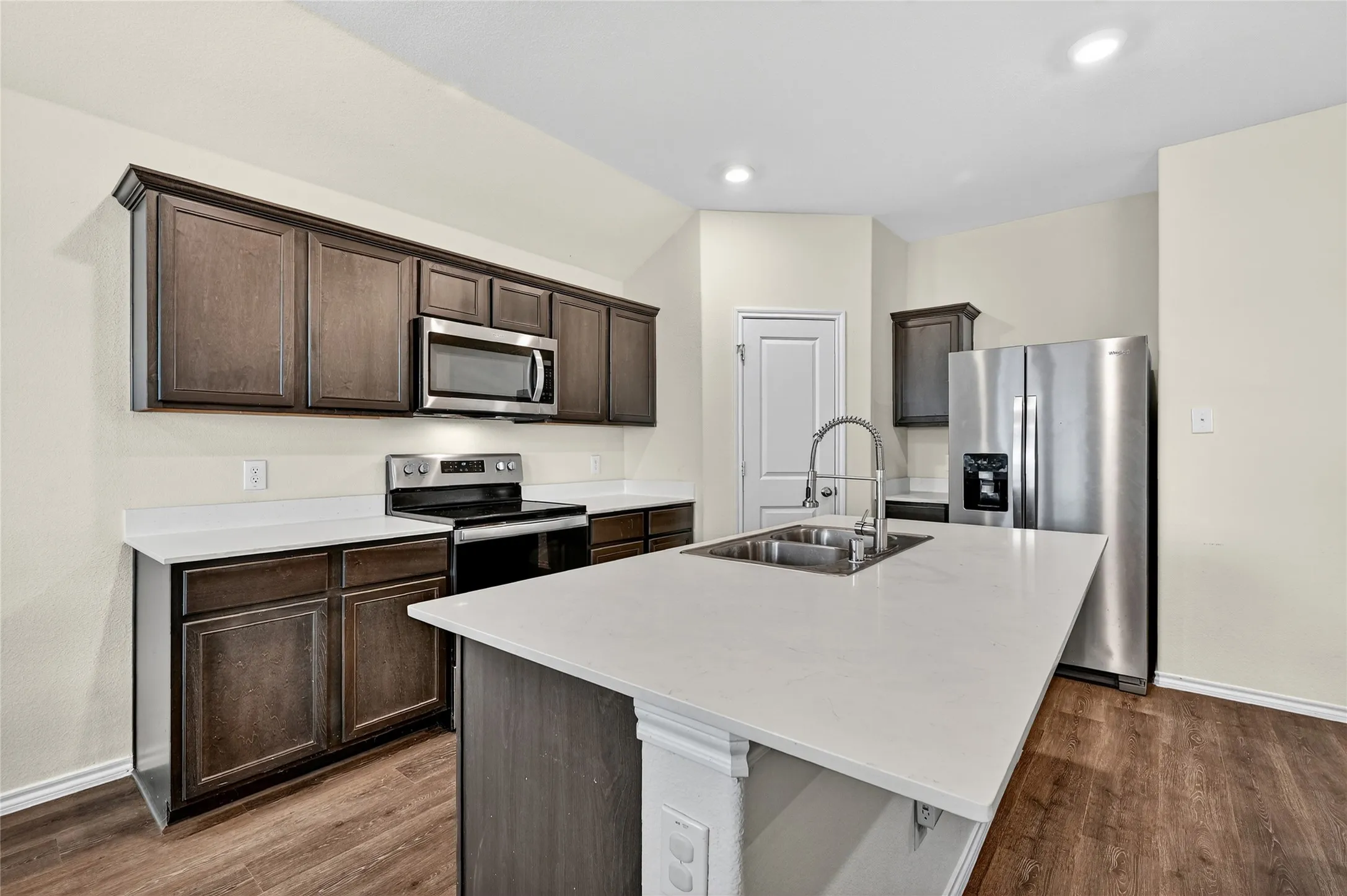 Kitchen featuring appliances with stainless steel finishes, dark brown cabinetry, an island with sink, dark wood-style flooring, and light countertops