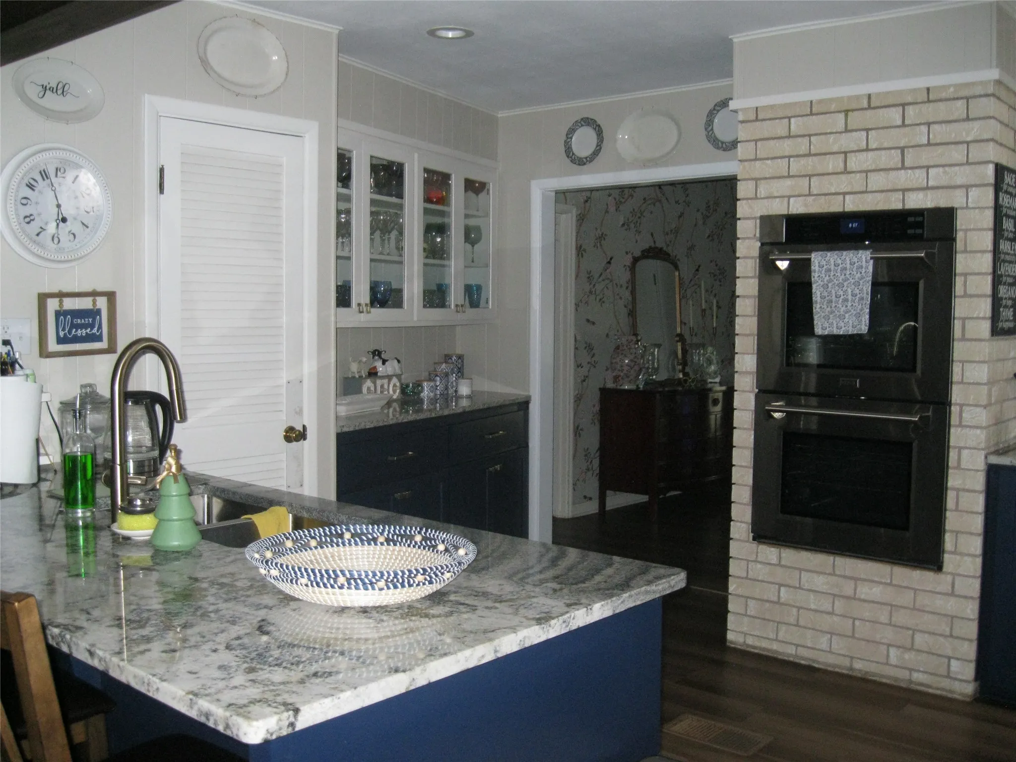 Kitchen with double wall oven, dark wood finished floors, crown molding, light stone countertops, and glass insert cabinets