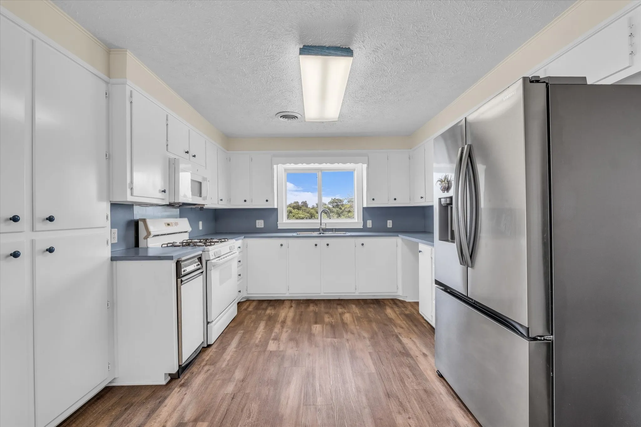 Kitchen featuring white appliances, a textured ceiling, wood finished floors, white cabinetry, and dark countertops