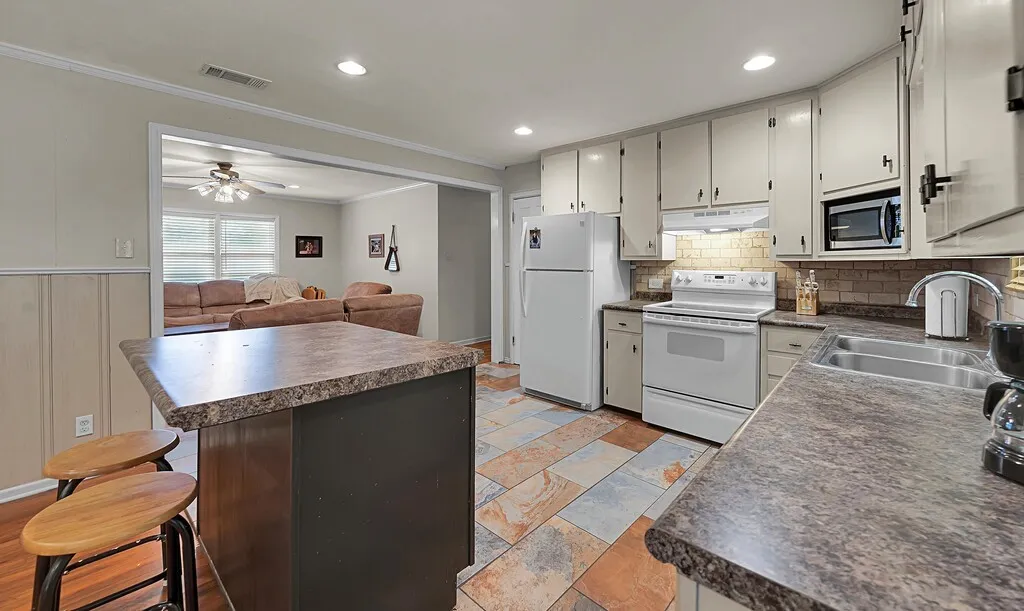 Kitchen with white appliances, under cabinet range hood, dark countertops, ornamental molding, and tasteful backsplash