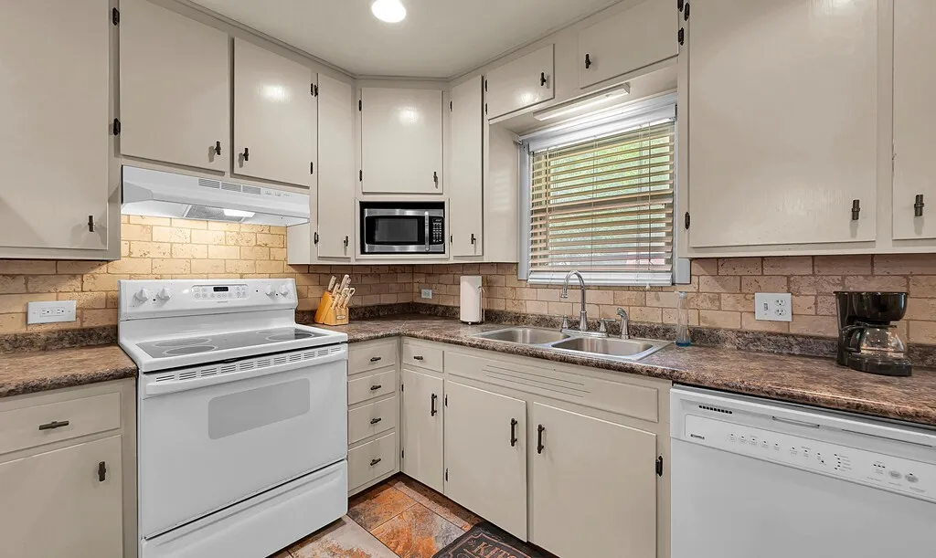 Kitchen featuring white appliances, under cabinet range hood, decorative backsplash, and dark countertops
