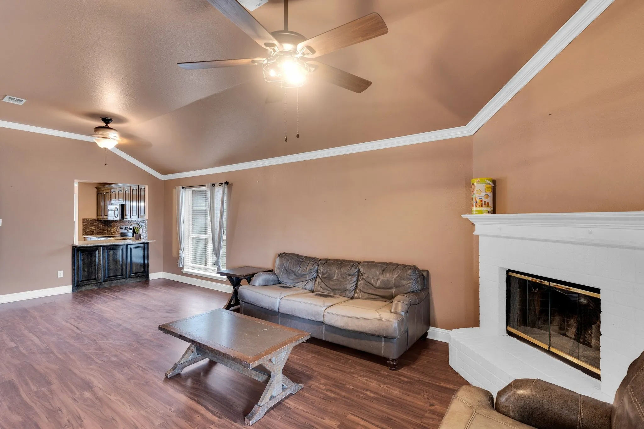 Living room with a ceiling fan, dark wood-type flooring, vaulted ceiling, a brick fireplace, and crown molding