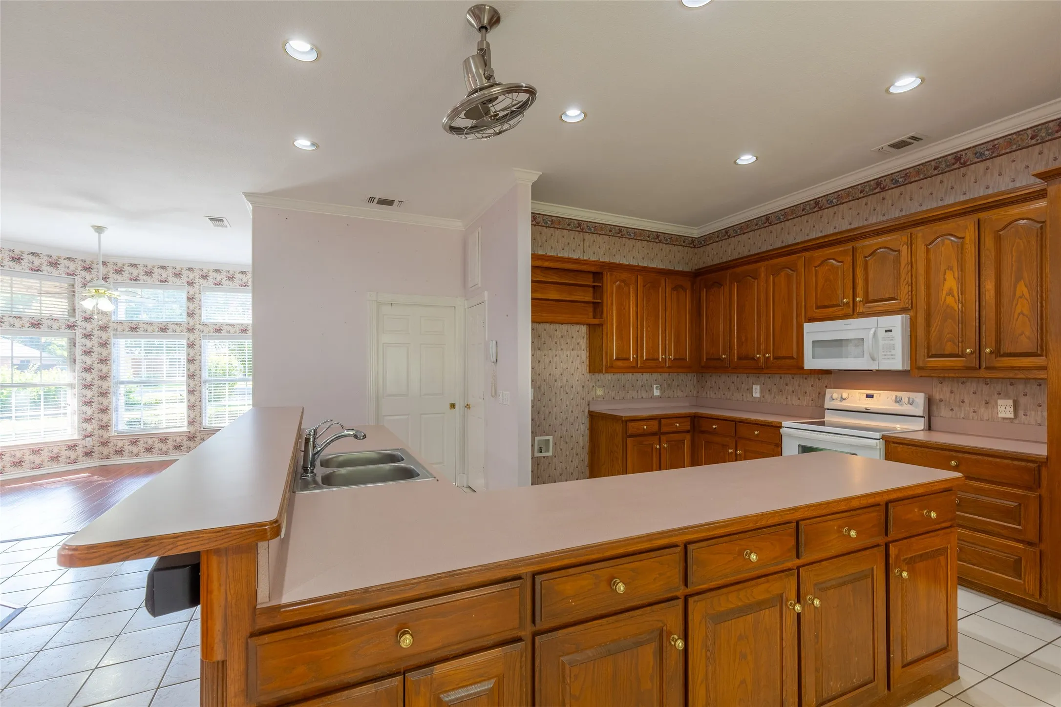 Kitchen featuring light tile patterned flooring, wallpapered walls, crown molding, brown cabinetry, and white appliances