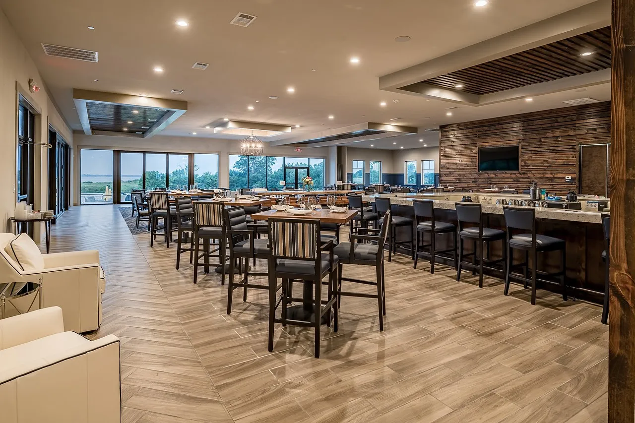Spacious interior featuring a large bar with a stone countertop and wood-paneled accent wall, a dining area with multiple tables, wood-look plank flooring in a herringbone pattern, recessed lighting, and expansive glass doors opening to the exterior