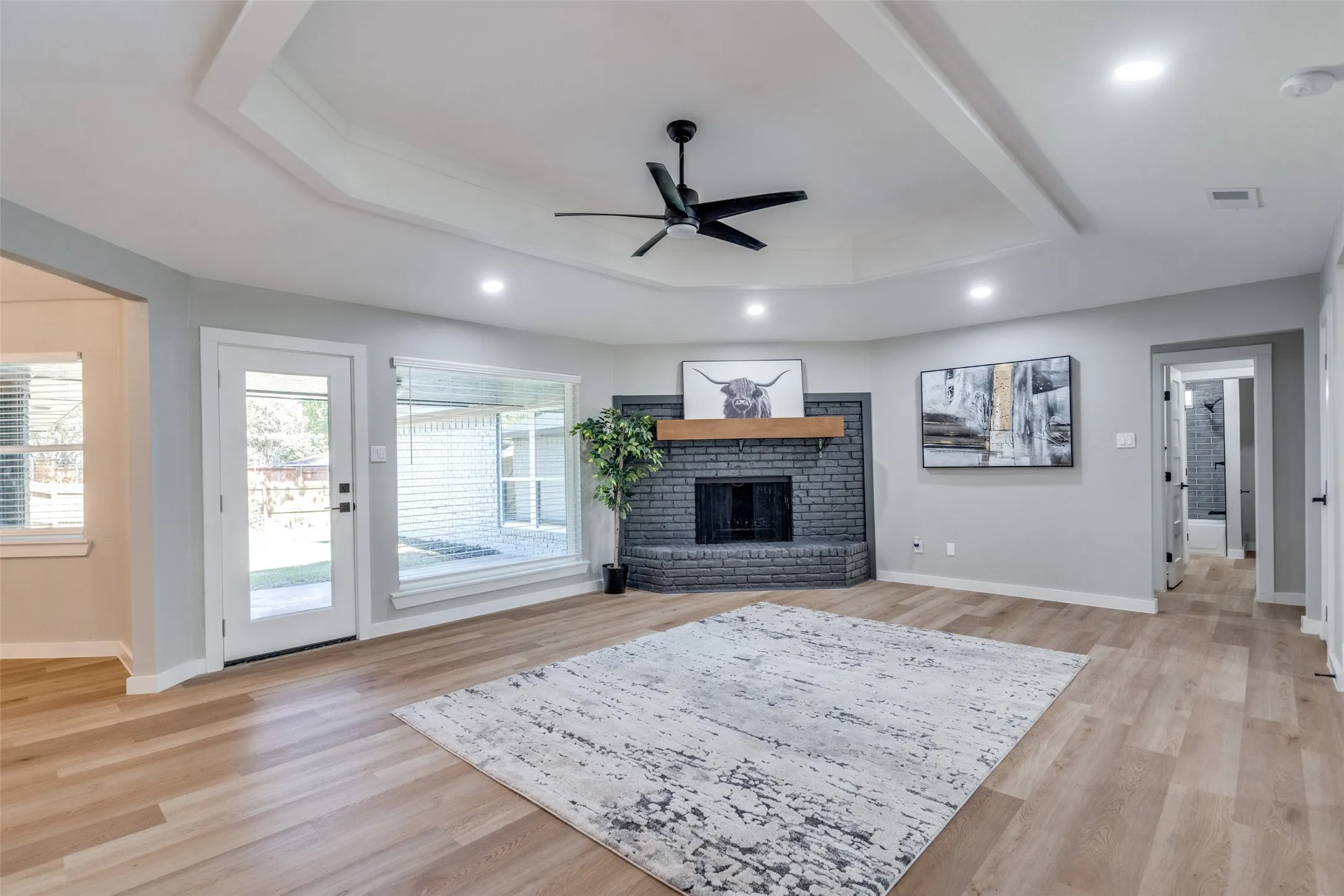 Living area featuring a tray ceiling, a brick fireplace, a ceiling fan, light wood-style flooring, and recessed lighting