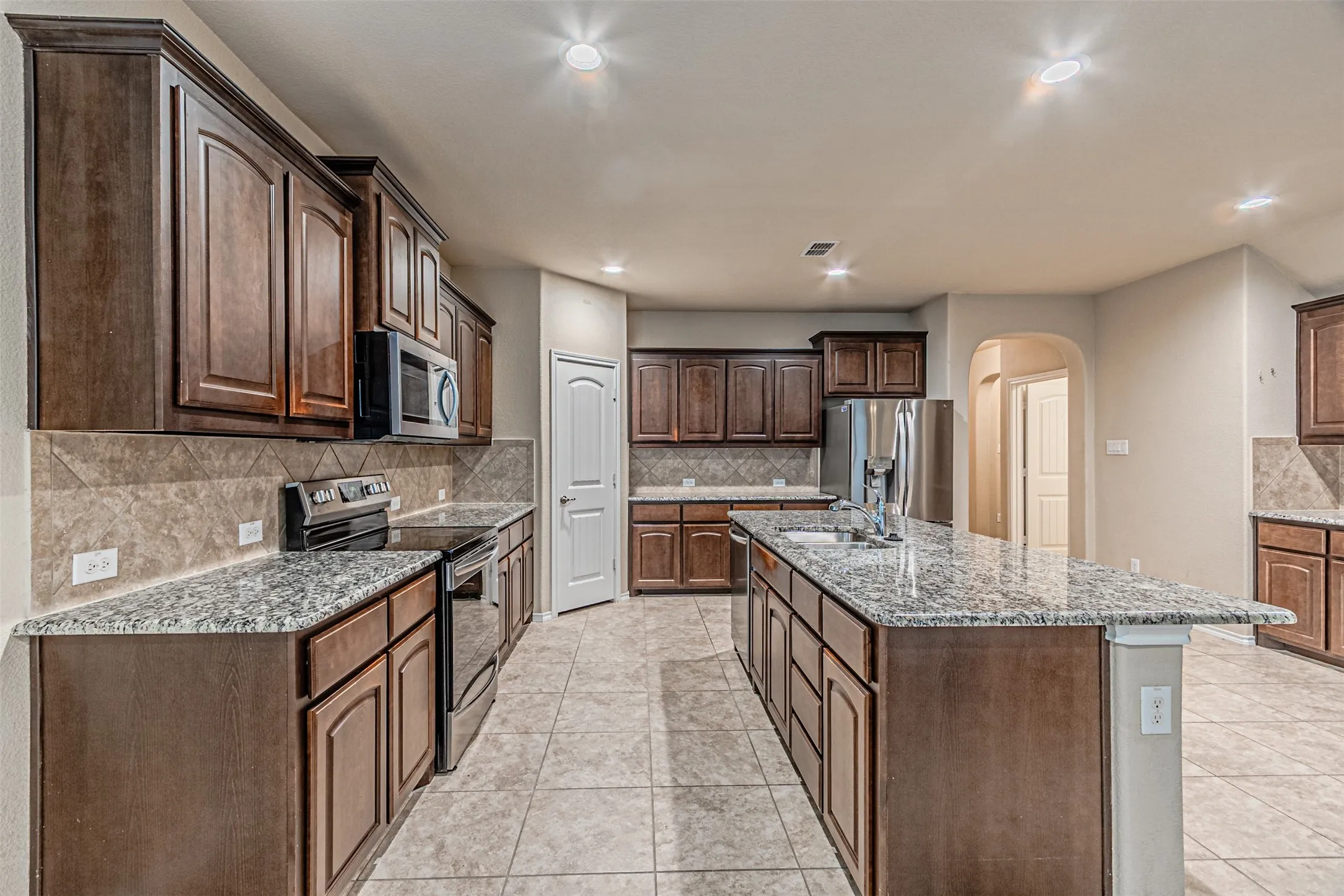 Kitchen with arched walkways, stainless steel appliances, tasteful backsplash, a kitchen island with sink, and light tile patterned flooring