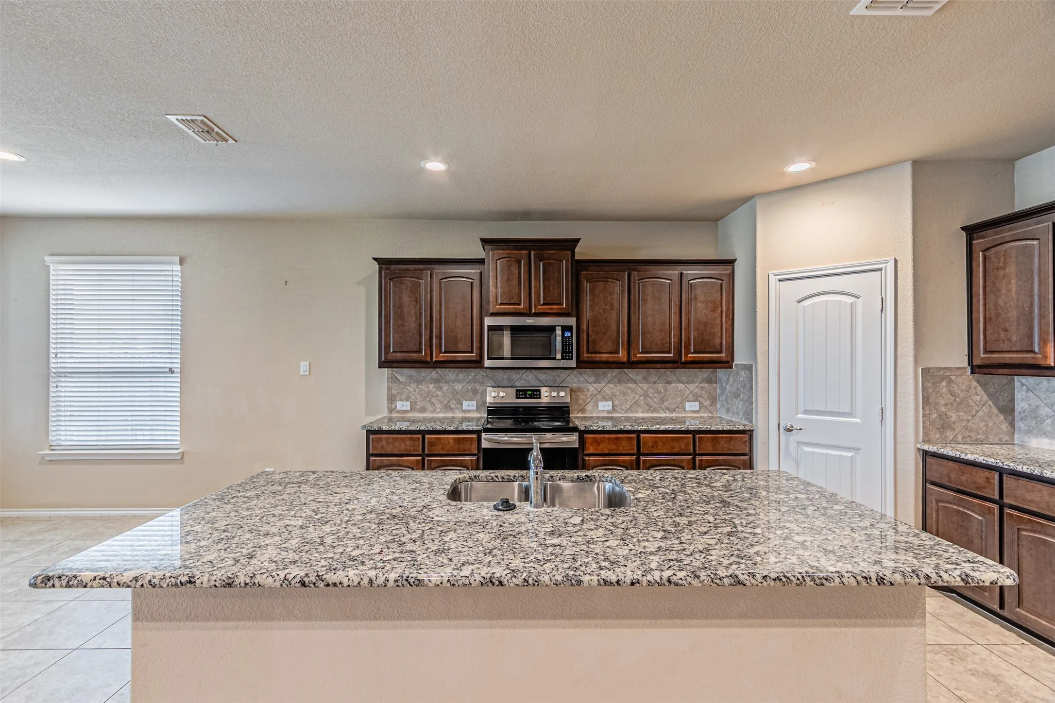 Kitchen featuring light tile patterned flooring, stainless steel appliances, a textured ceiling, light stone counters, and a kitchen island with sink