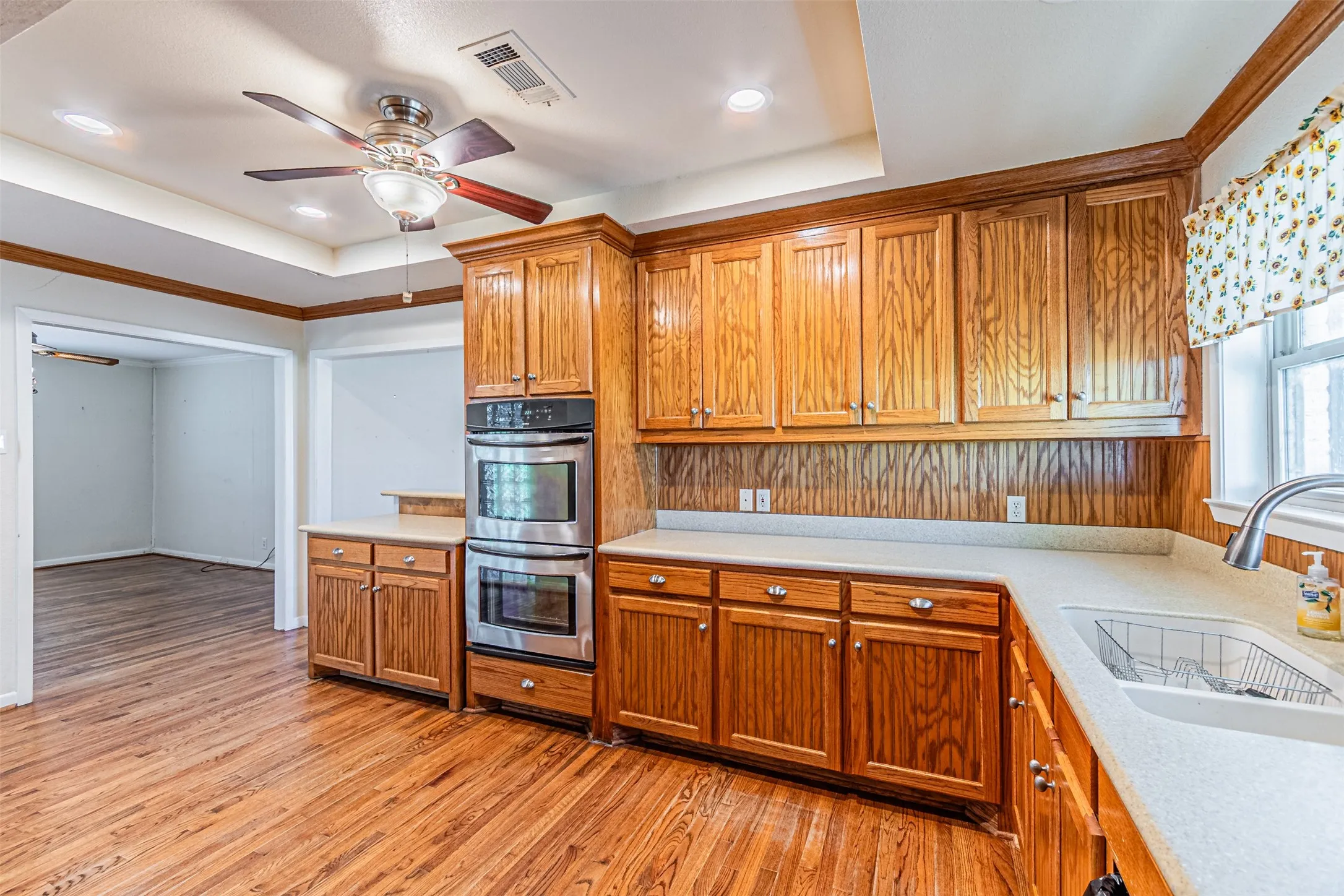 Kitchen with a raised ceiling, brown cabinetry, a ceiling fan, recessed lighting, and crown molding