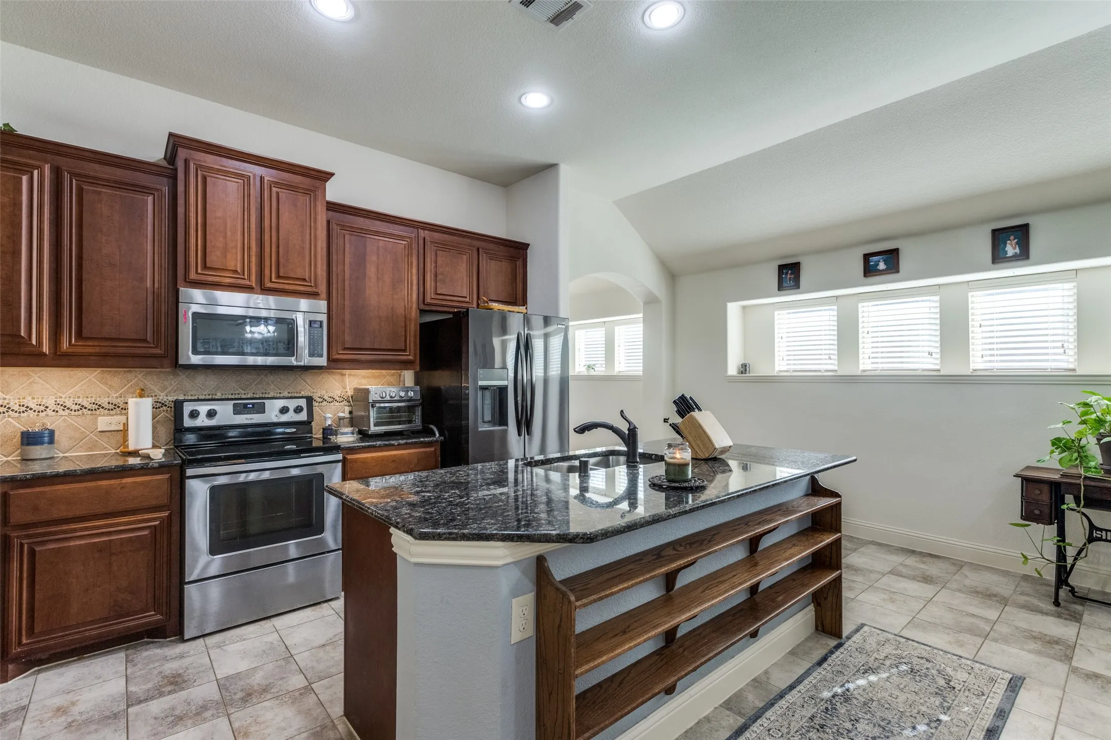 Kitchen with decorative backsplash, dark stone countertops, stainless steel appliances, arched walkways, and a center island with sink