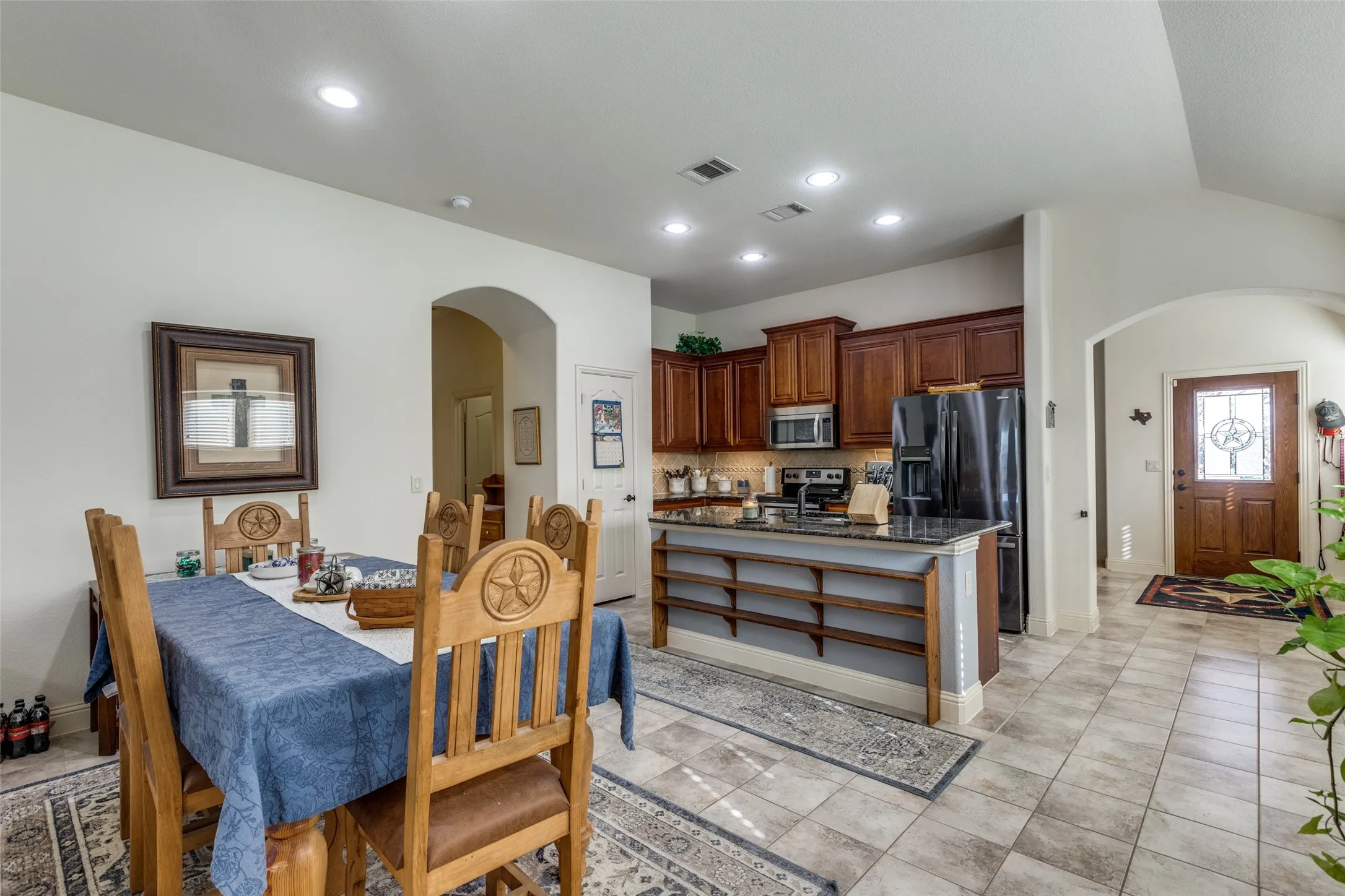 Kitchen featuring arched walkways, backsplash, an island with sink, dark stone countertops, and open shelves