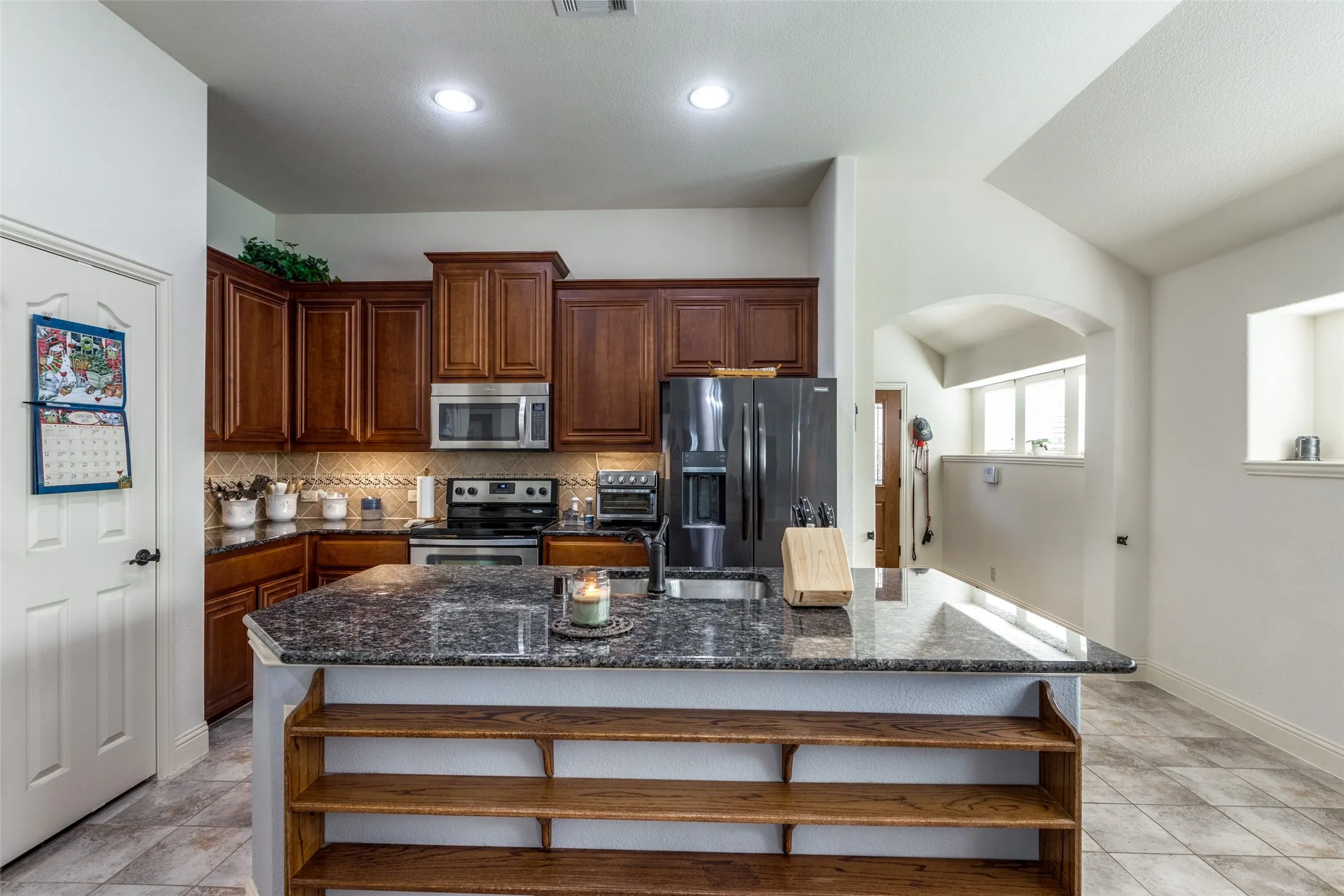 Kitchen with dark stone counters, open shelves, backsplash, stainless steel appliances, and an island with sink