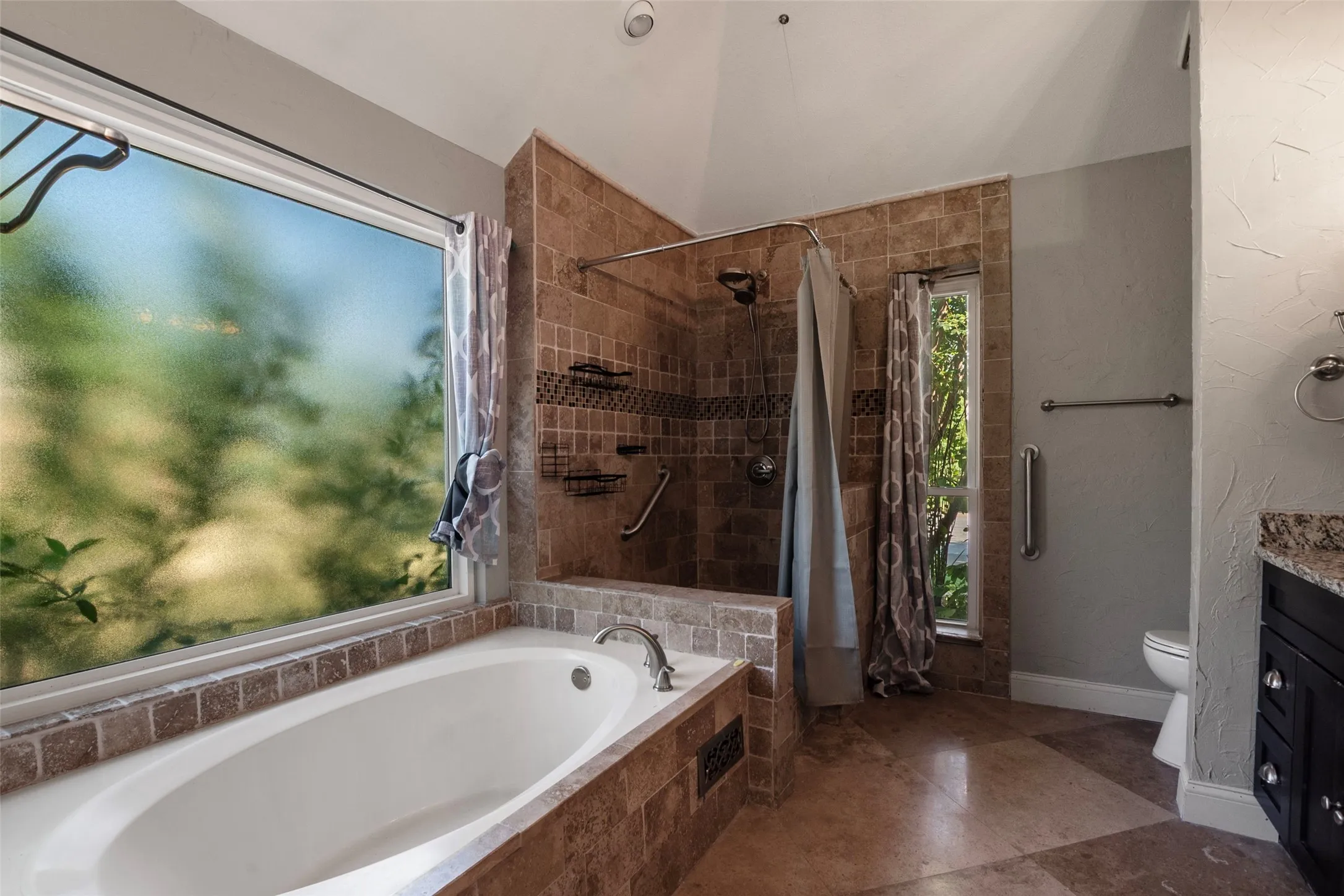 Soaking tub with a tiled surround, tiled walk-in shower with a glass enclosure, vaulted ceiling, and a vanity with a stone countertop