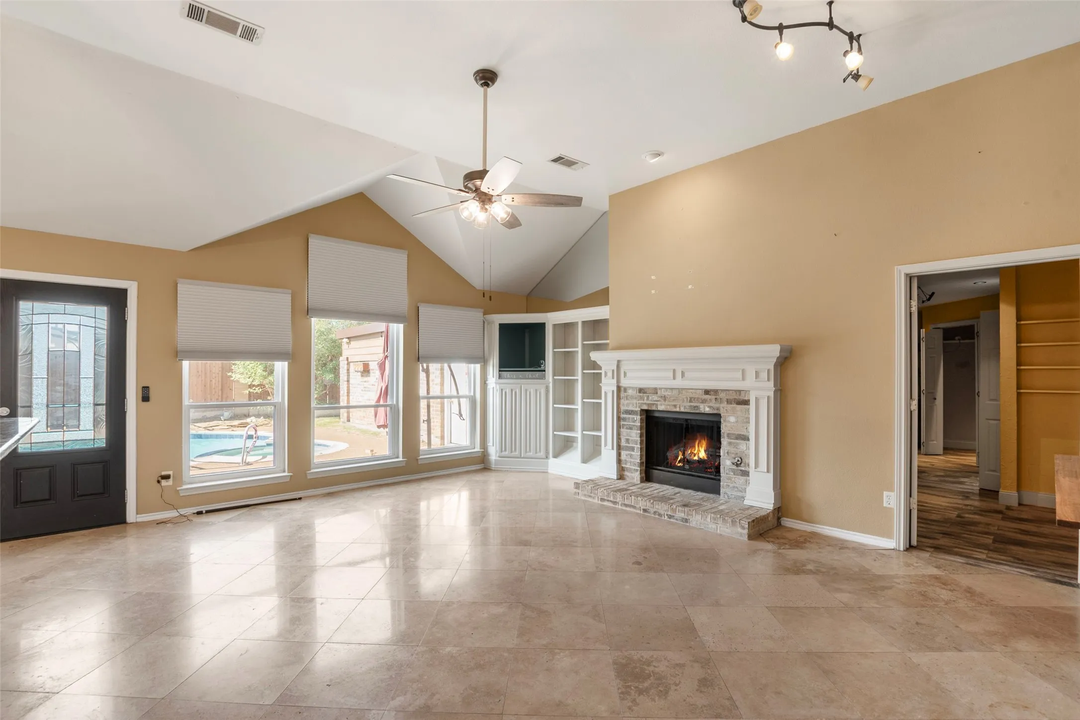 Vaulted ceilings, large format tile flooring, brick fireplace with mantel, built-in shelving, and multiple windows overlooking a backyard pool