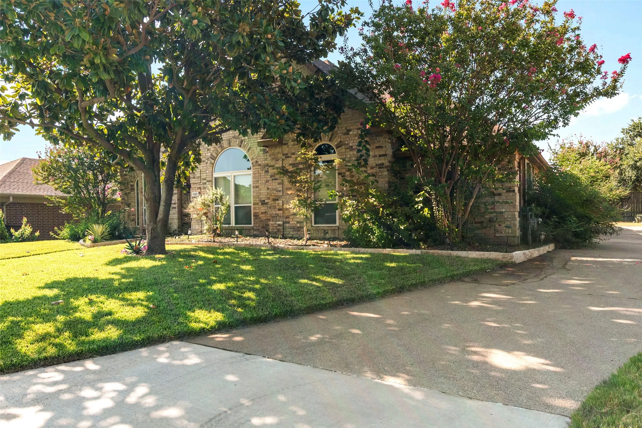 Brick exterior with arched window, concrete driveway, front lawn, and mature trees with foliage
