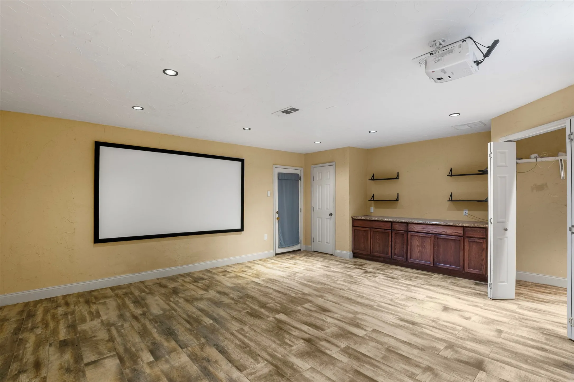Spacious room with wood-finish flooring, recessed lighting, built-in media screen, ceiling-mounted projector, and a built-in wood cabinet with shelving