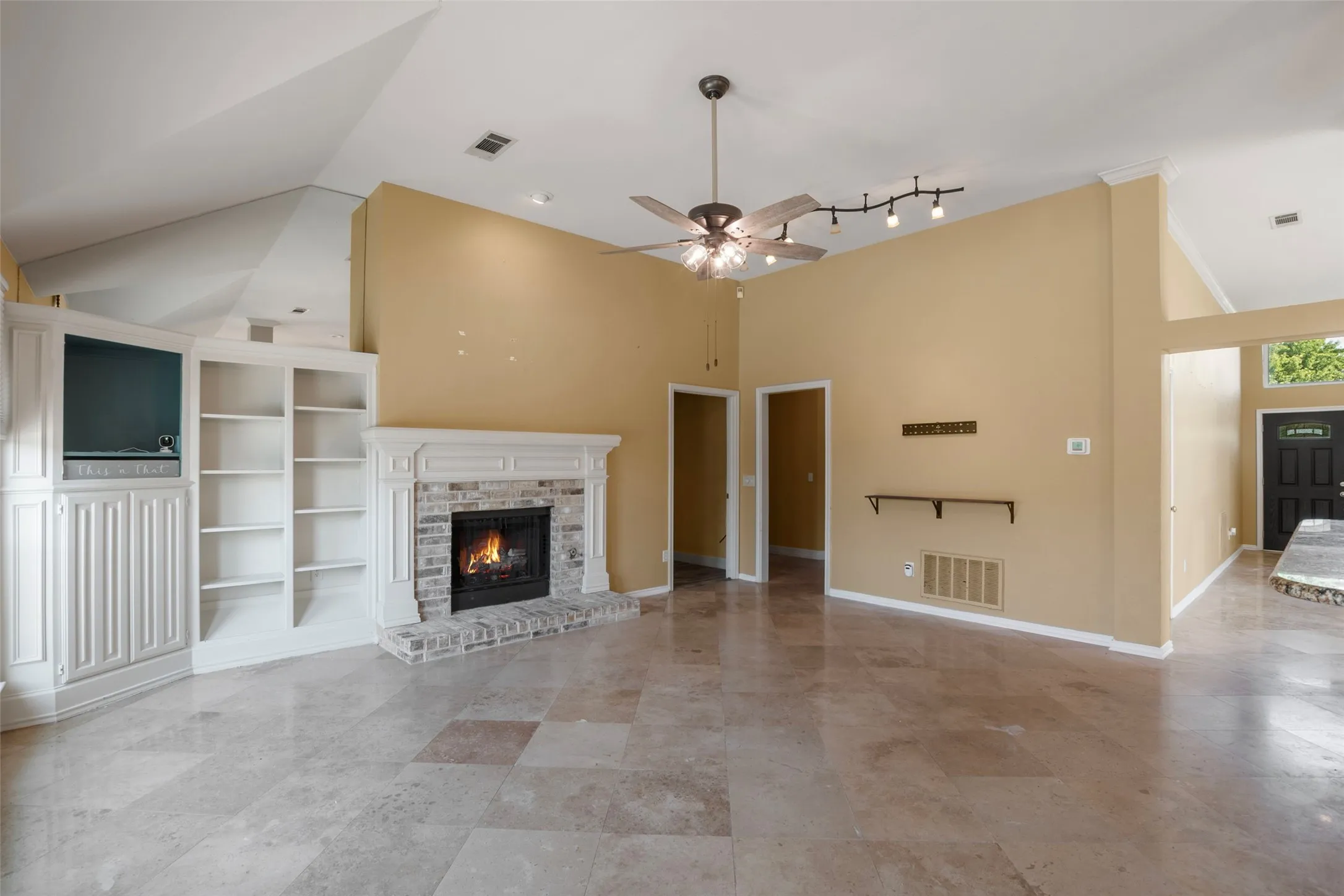 Vaulted ceiling with an exposed beam, a stone fireplace with a white mantel, built-in shelving, a ceiling fan, and tile flooring