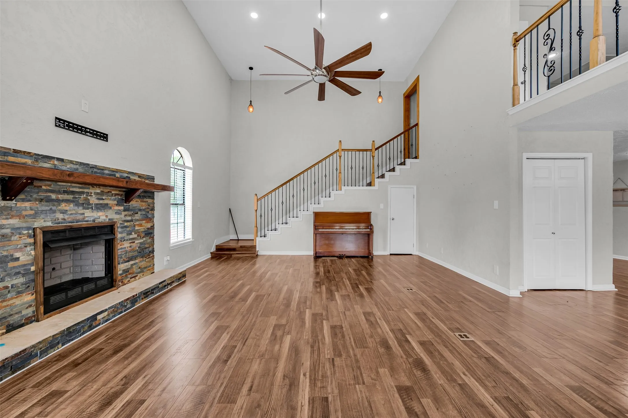 Unfurnished living room with a stone fireplace, high vaulted ceiling, wood finished floors, ceiling fan, and stairway