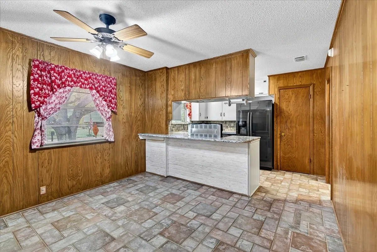 Kitchen featuring wooden walls, a peninsula, black fridge, a ceiling fan, and light stone countertops