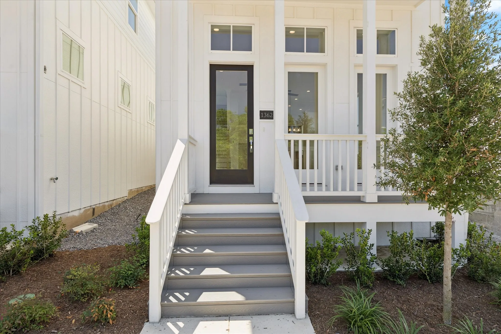 Inviting front entry featuring a dark-framed glass door, white board and batten siding, a covered porch with white railings, and a mature tree
