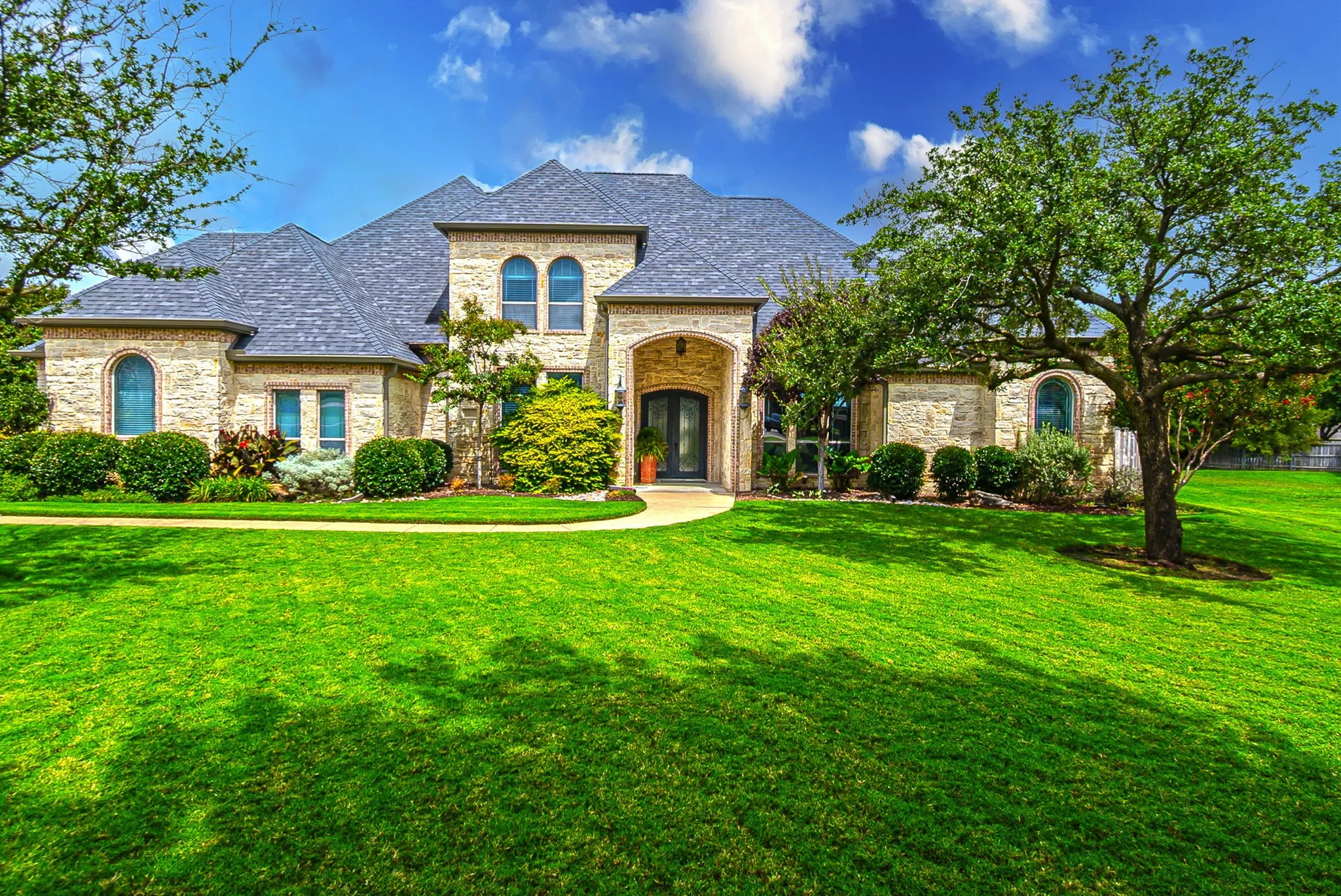 Front of home featuring large yard, mature trees, double iron front doors with glass inserts, and beautiful landscaping.