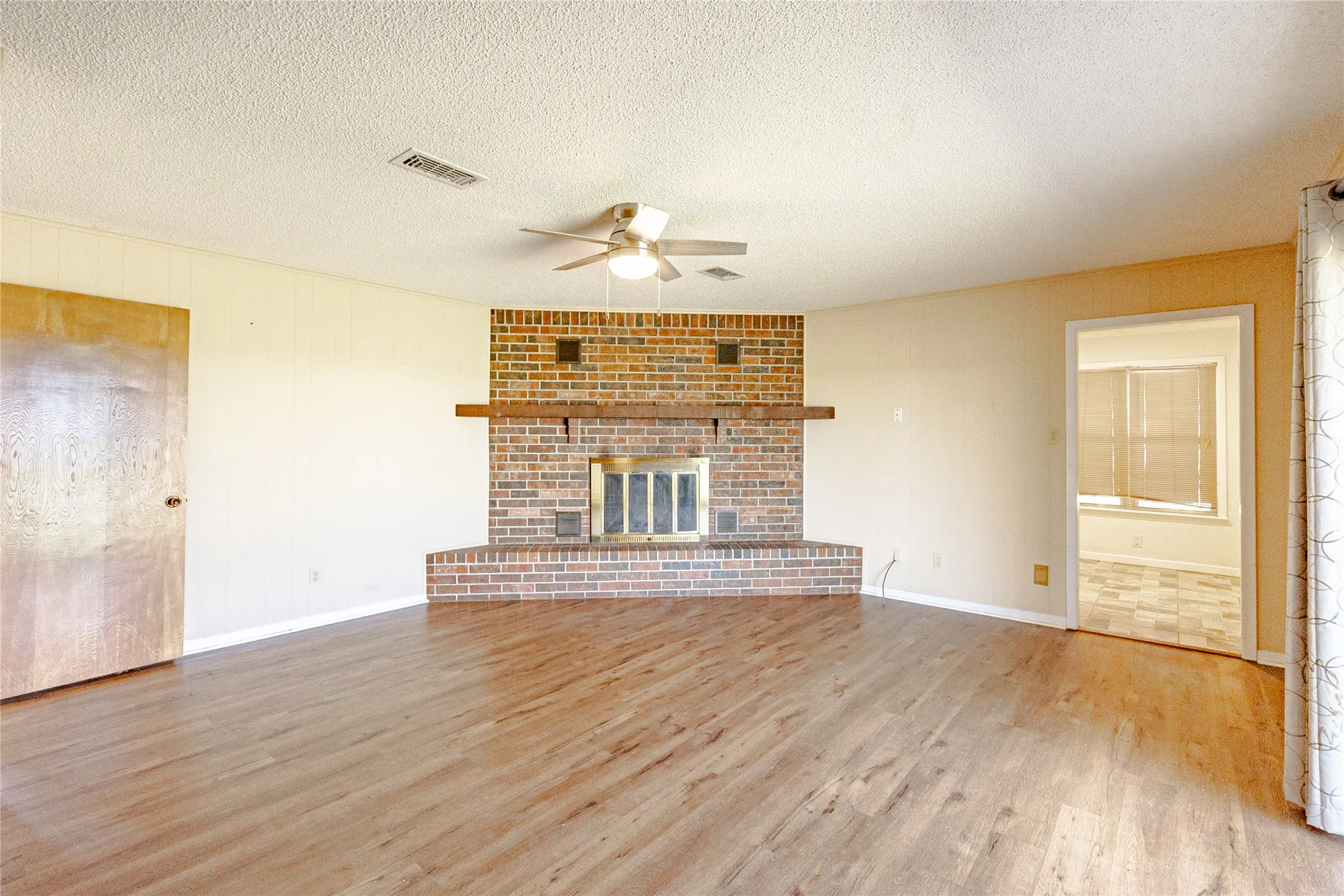 Unfurnished living room with light wood-style floors, a brick fireplace, a ceiling fan, a textured ceiling, and wooden walls
