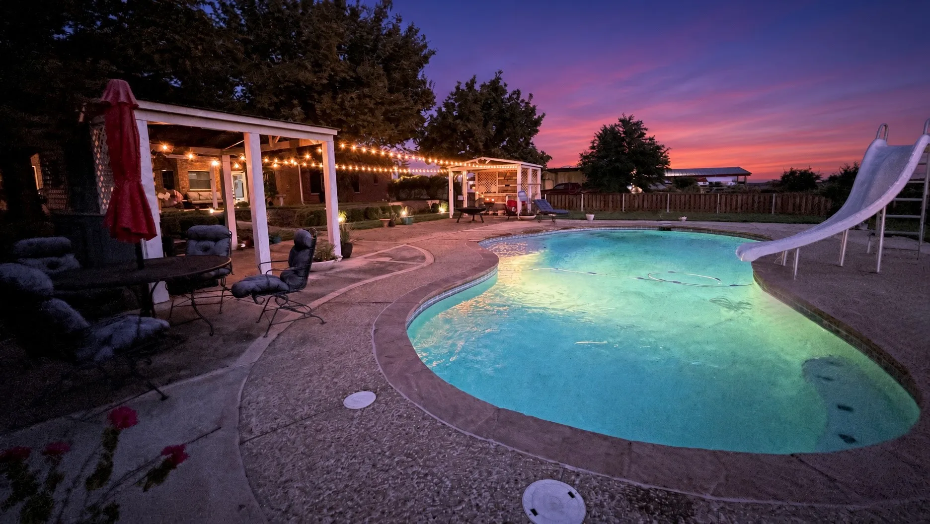 Freeform swimming pool with integrated slide, surrounded by a textured concrete deck, and an adjacent open-air pergola